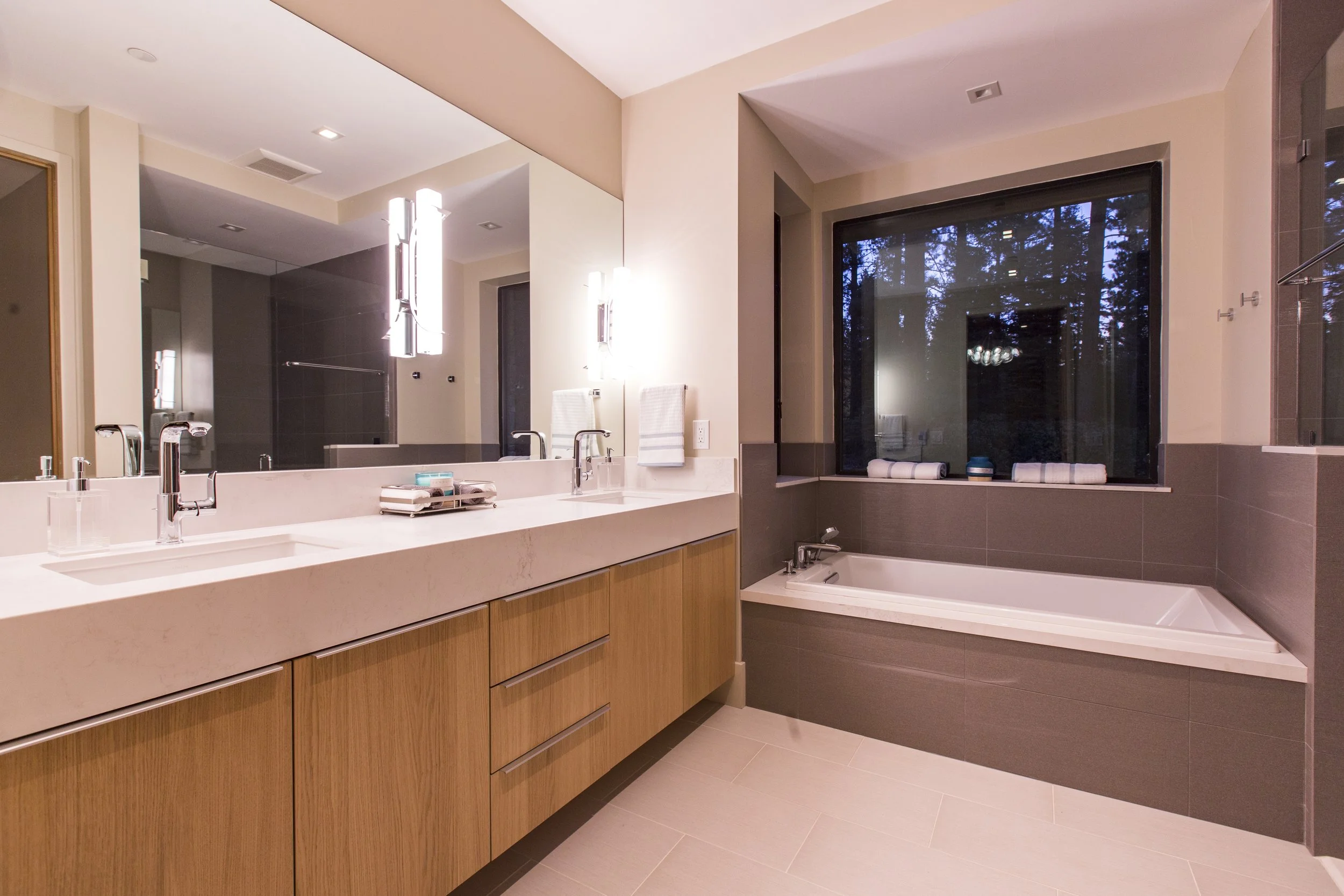 Modern bathroom with double sink vanity, large mirror, and a bathtub next to a window with a view of trees outside.