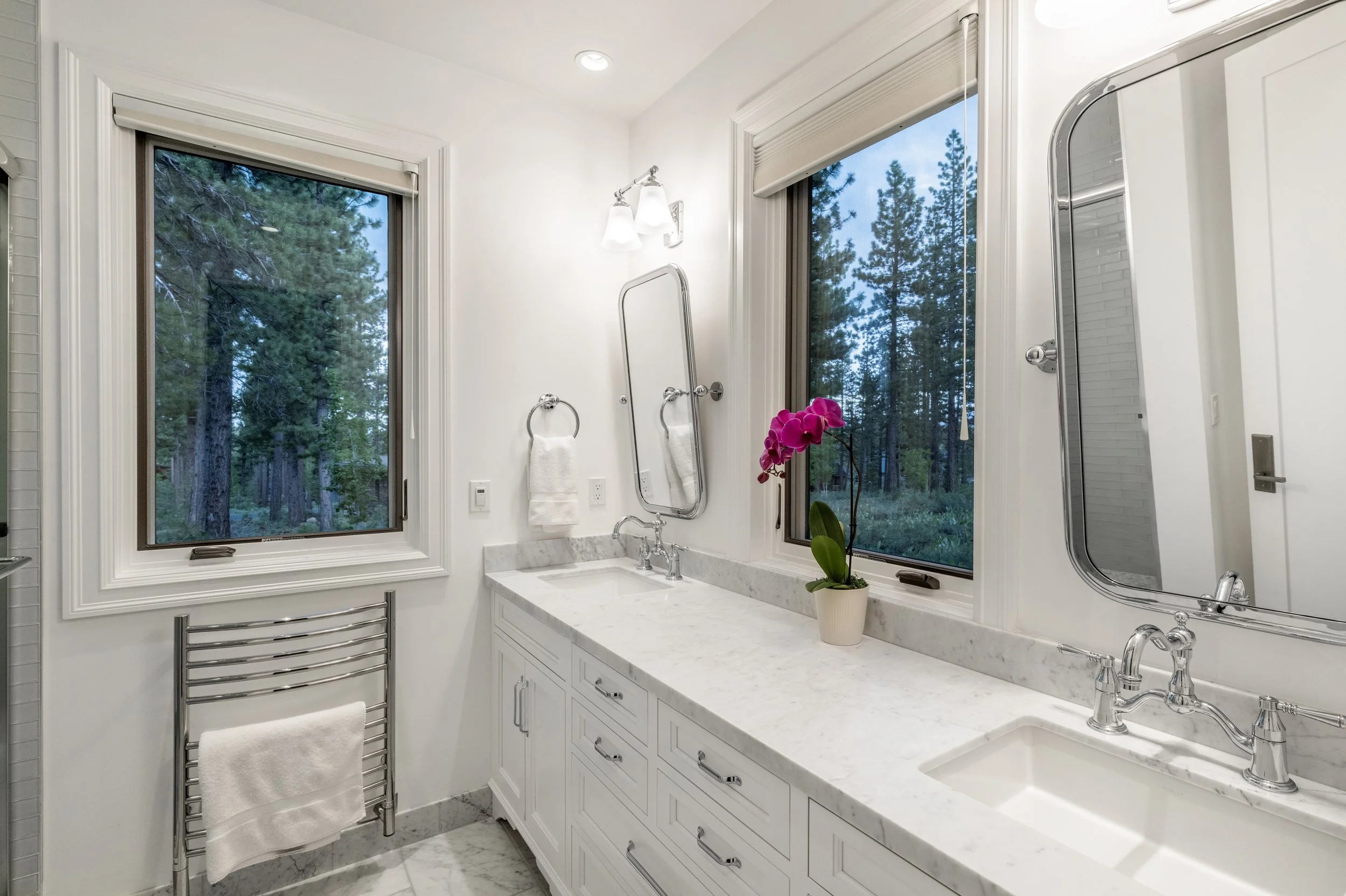 A bright bathroom with two large windows showing trees outside, white cabinetry, marble countertops, a potted orchid on the counter, and two mirrors above the sinks.