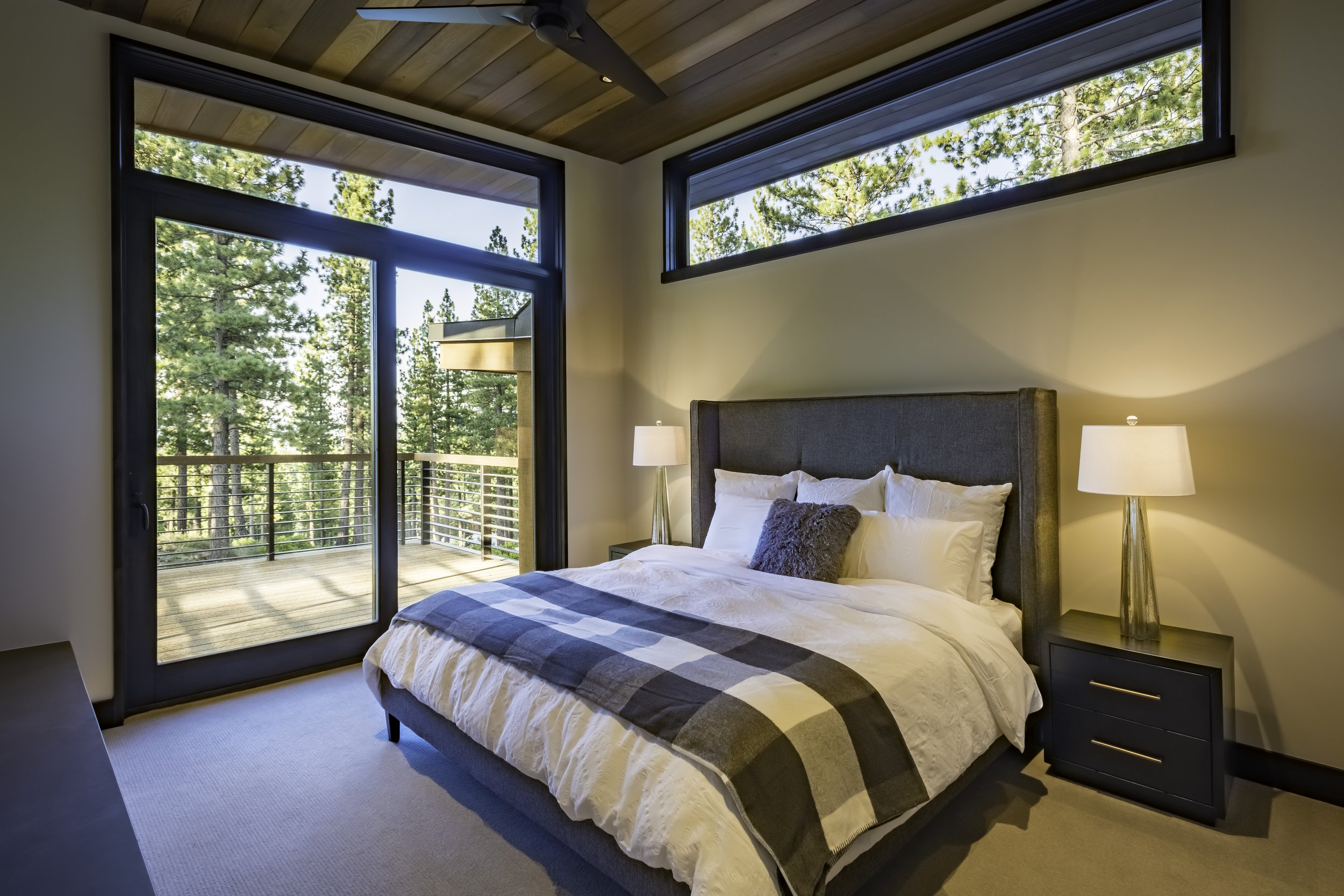 A bedroom with a bed, two nightstands with lamps, a sliding glass door opening to a balcony, and large horizontal windows showing trees outside.