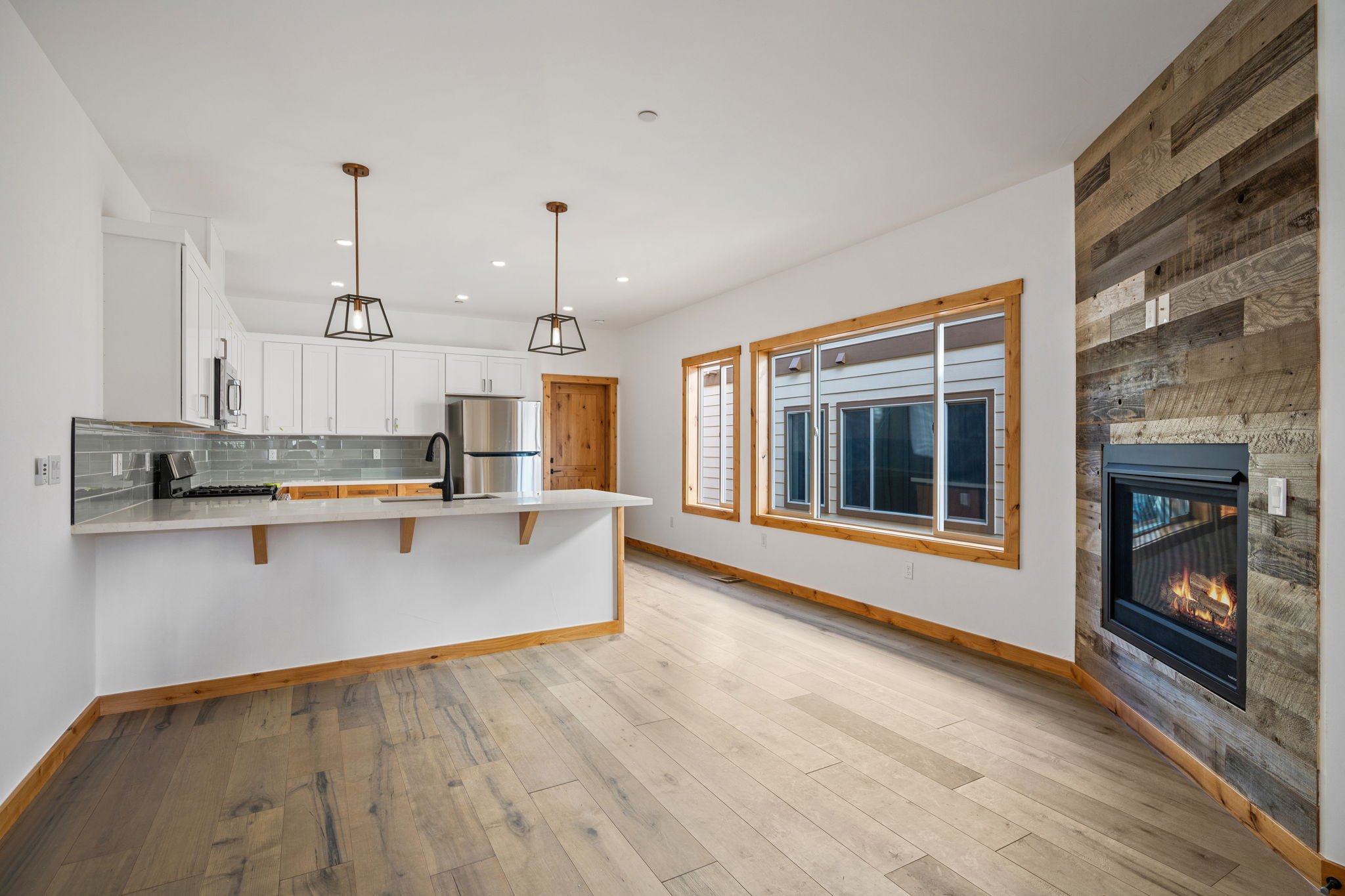 Open kitchen and living area with white cabinets, stainless steel refrigerator, black sink, wood accents, with large windows and a modern fireplace with wood paneling on the wall.
