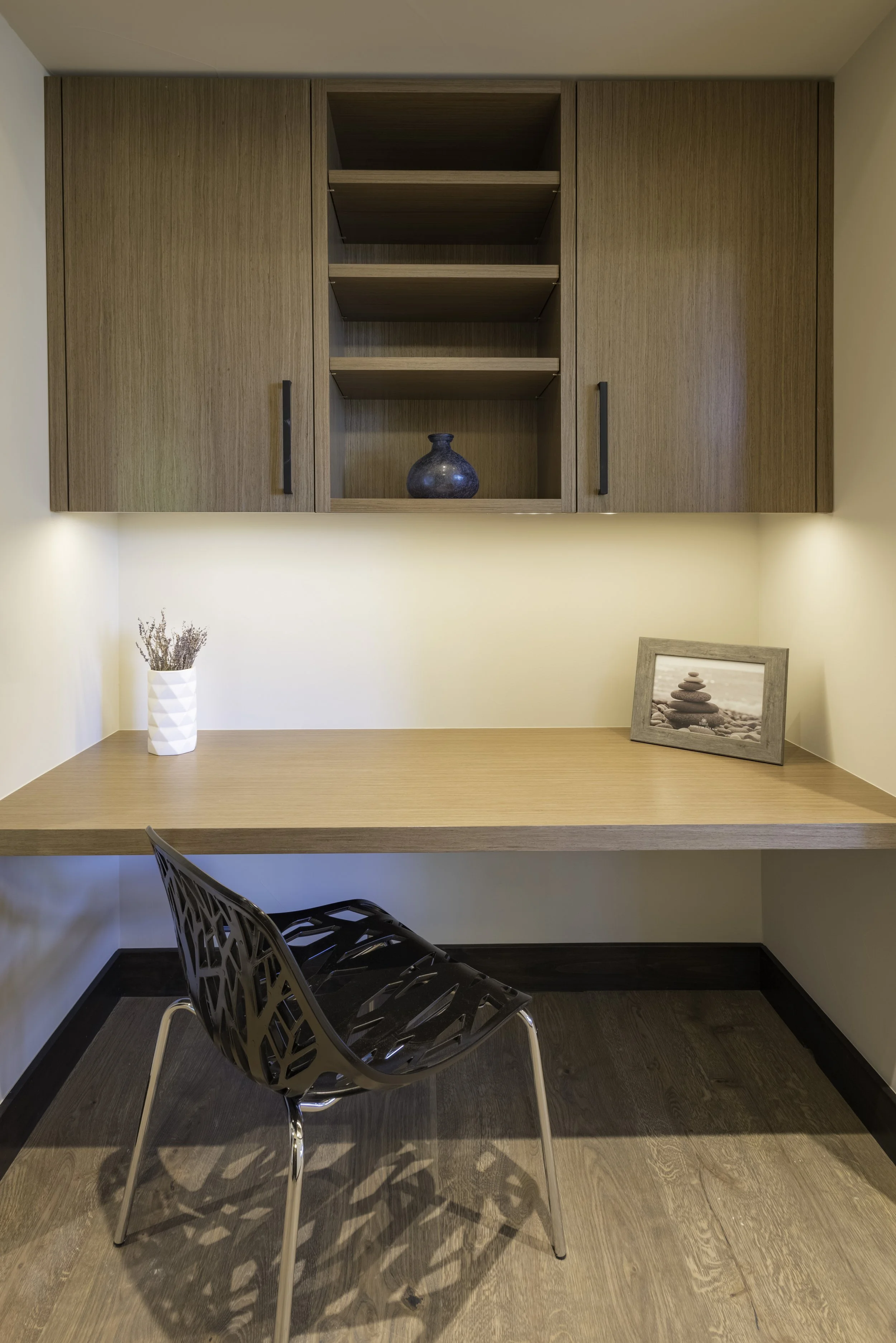 Small home office with a wooden desk, black modern chair, native decor and wall-mounted cabinets with open shelving and cabinet doors, illuminated with subtle lighting.