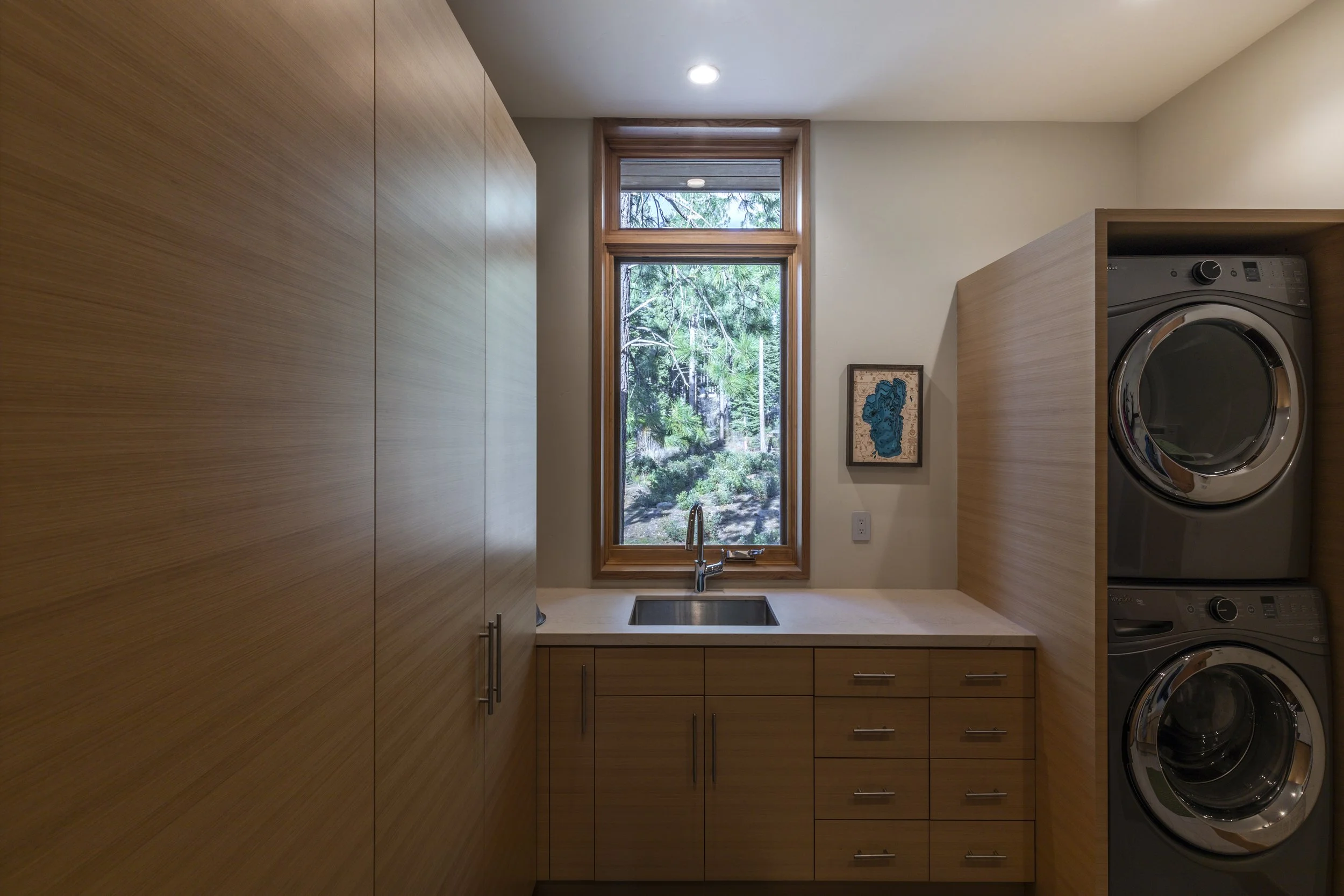 Laundry room with wooden cabinets, a window overlooking trees, a white countertop with a sink, and stacked washer and dryer machines.