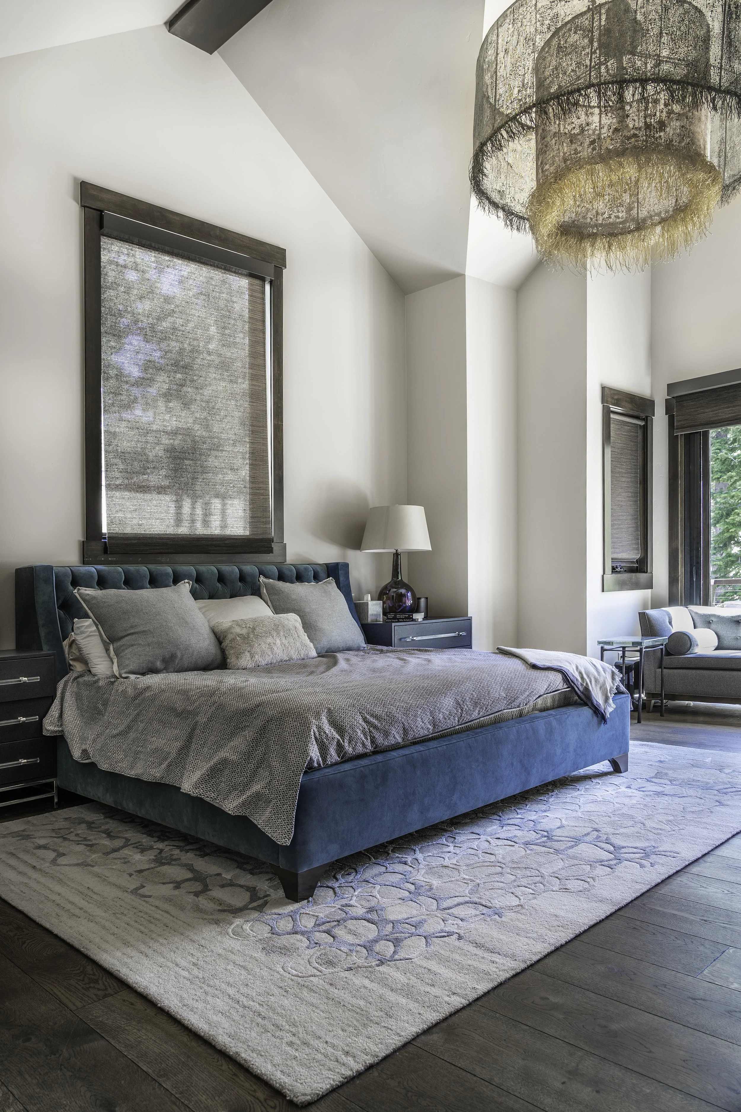 A modern bedroom featuring a teal upholstered bed, gray bedding and pillows, dark wood nightstands, a bedside lamp, a white area rug, and windows with brown blinds. There is a textured chandelier hanging from the ceiling.