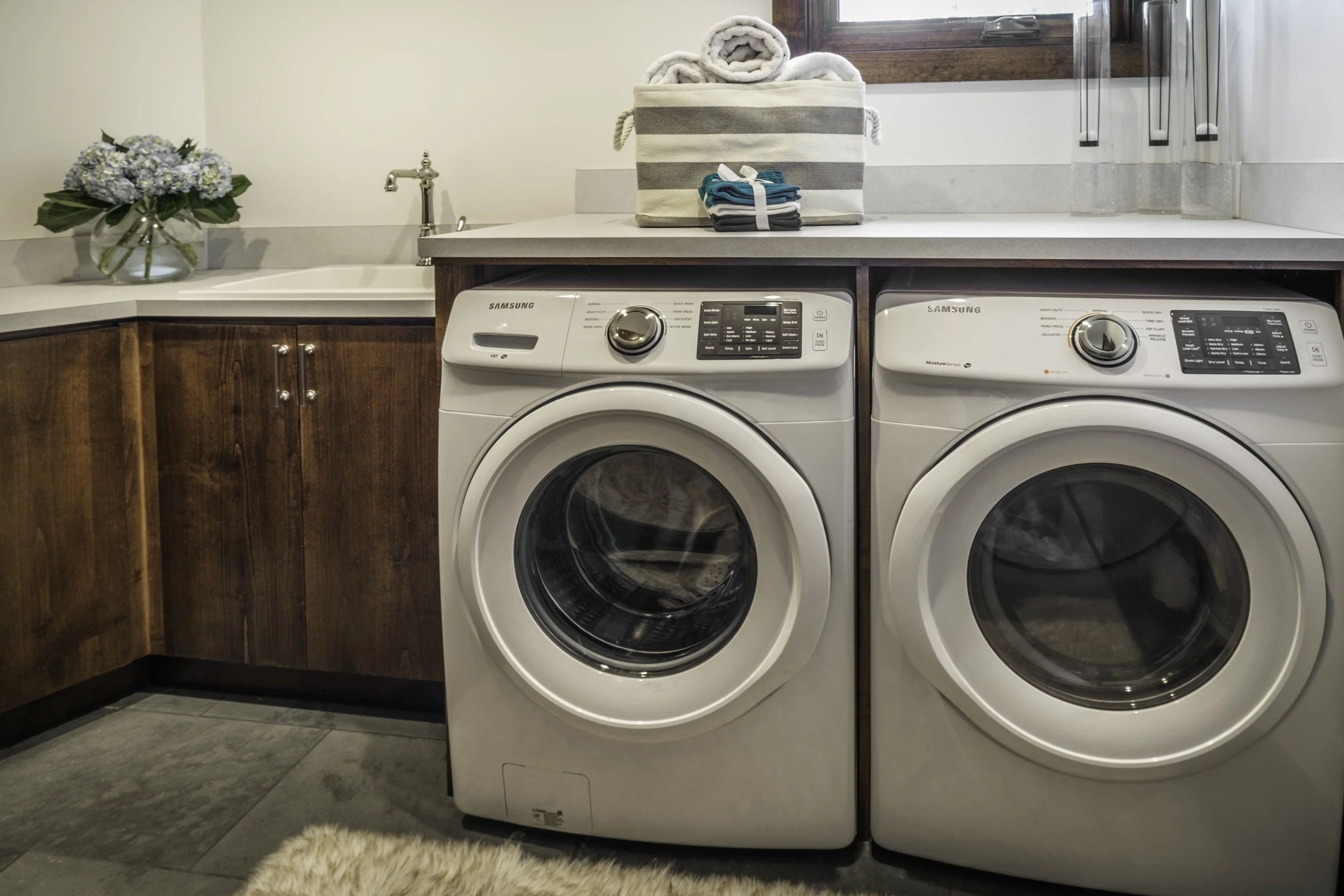 Front-loading washing machine and dryer in laundry room with a wooden cabinet, a vase of hydrangeas, and folded towels on countertop.
