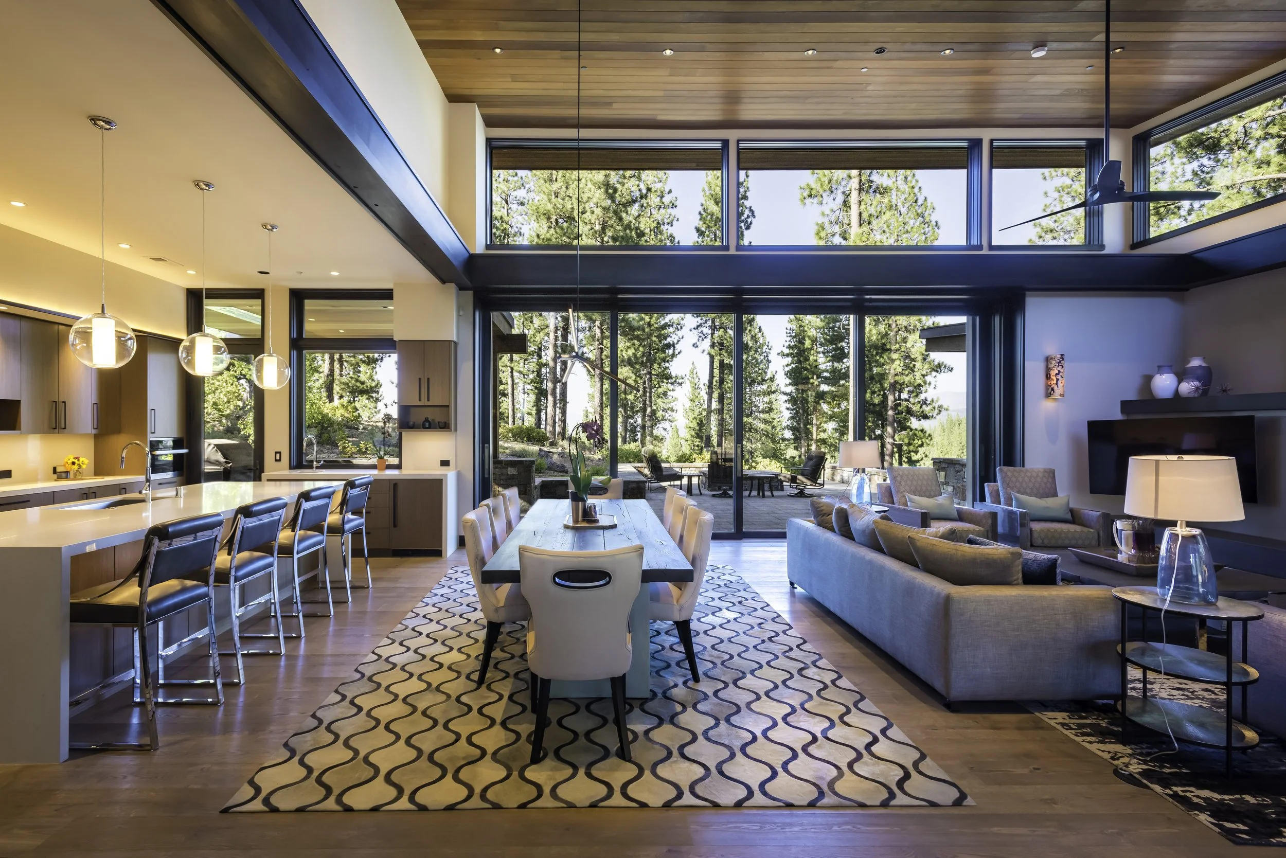 Modern open-concept living space with large floor-to-ceiling windows overlooking a wooded outdoor patio, featuring a kitchen, dining area, and living room with sofas, chairs, and lamps.