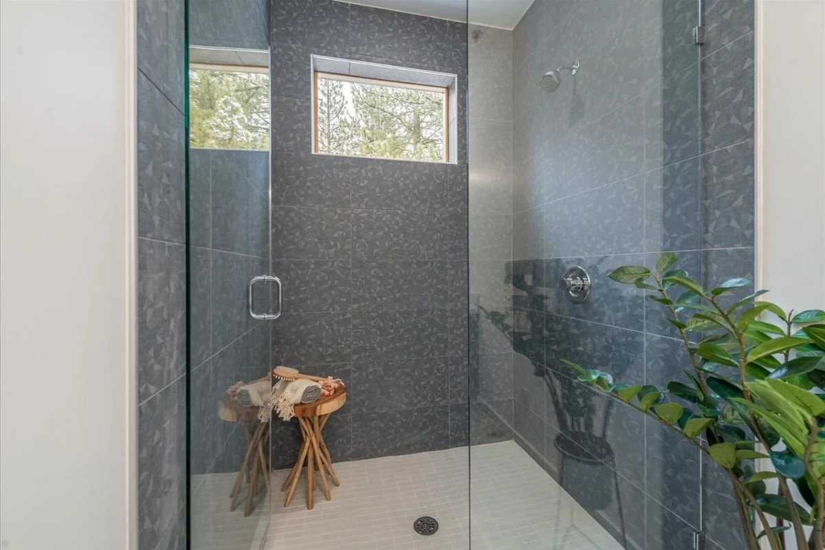 Contemporary large walk-in shower with dark gray textured tiles on the walls, a small horizontal window, and a glass door, with a wooden stool and green plant outside the shower.