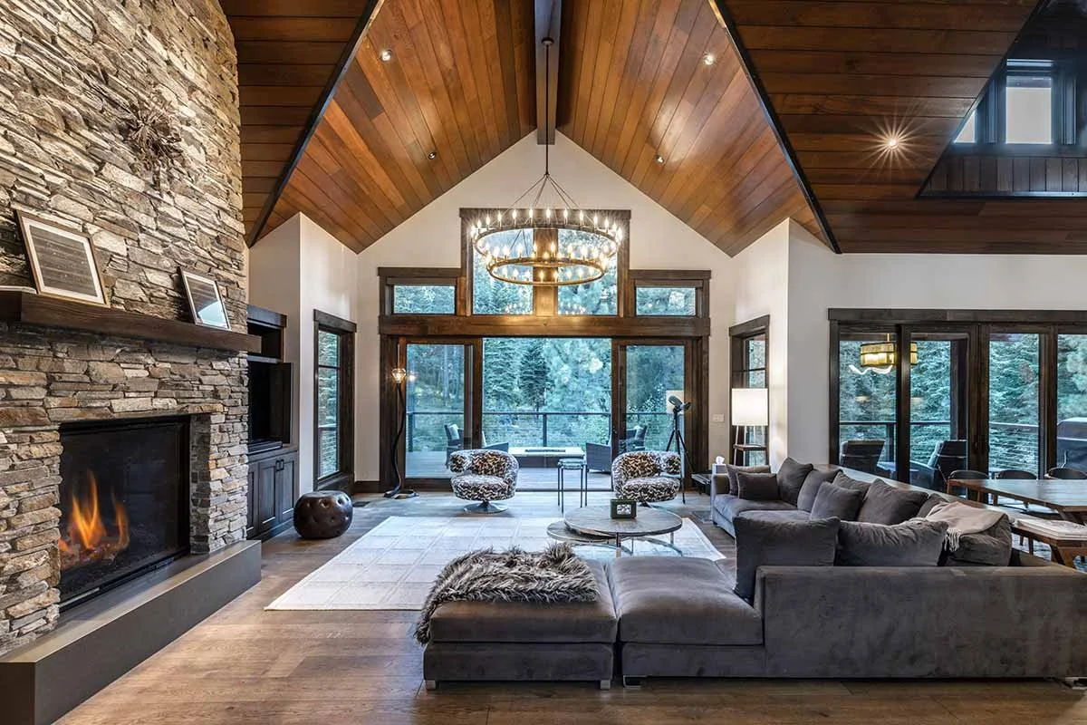 Spacious living room with a vaulted wooden ceiling, large stone fireplace, modern chandelier, and furniture including sofas, chairs, and a coffee table, with large windows and a balcony overlooking trees.