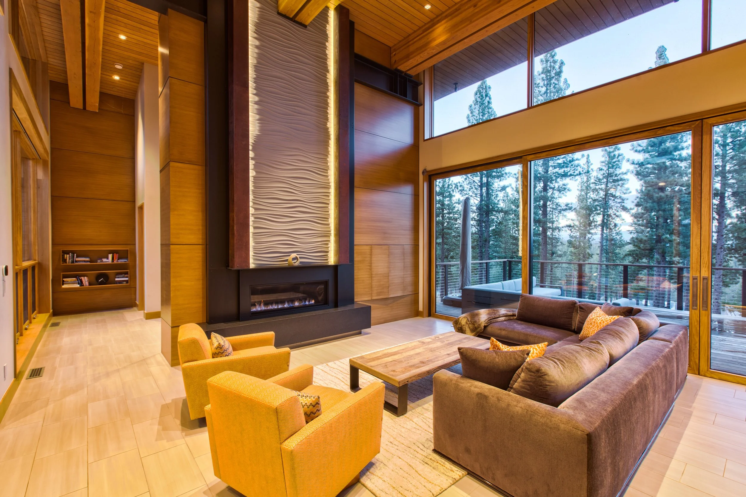 Living room with large floor-to-ceiling windows showing trees outside, a brown sofa, yellow armchairs, a wooden coffee table, a modern fireplace, and a textured wall panel above the fireplace.