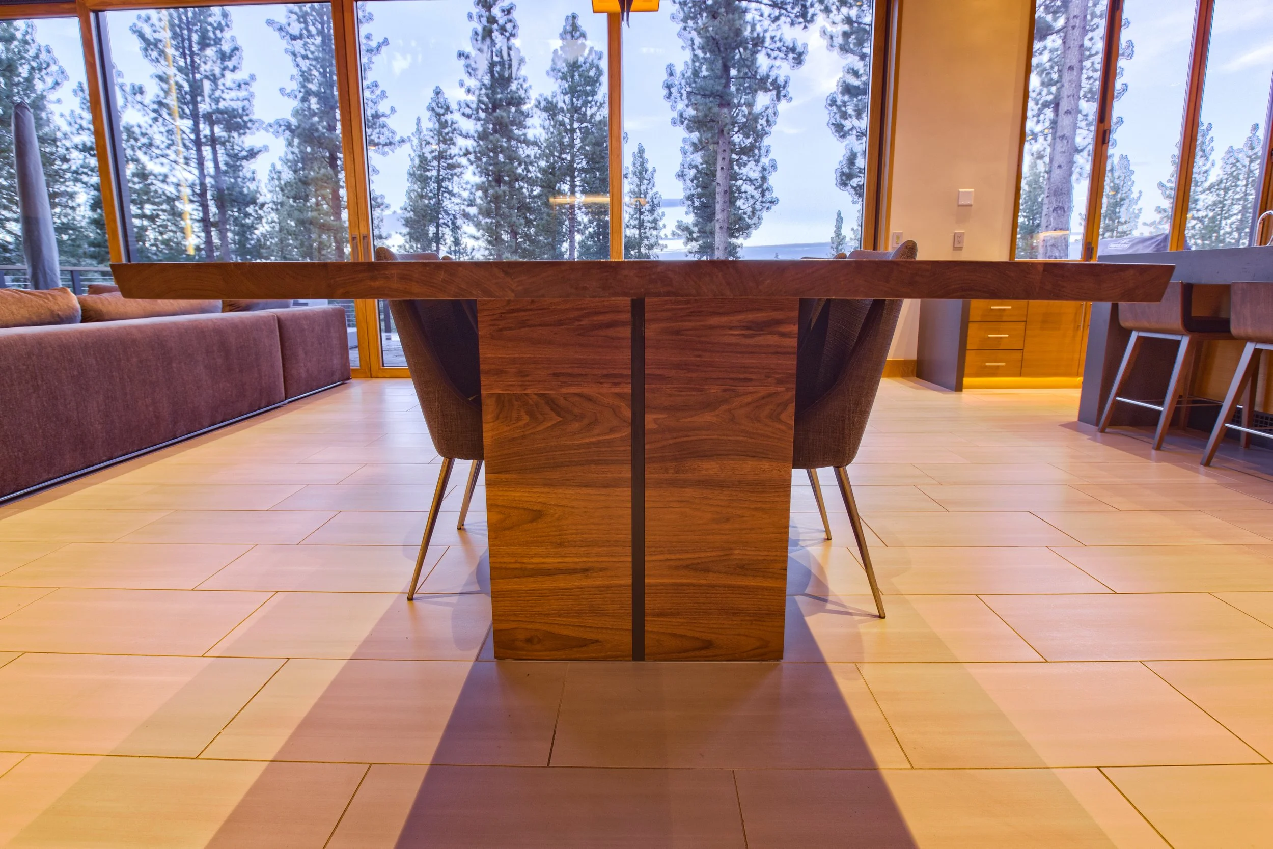 Interior view of a dining room with a large wooden table, gray chairs, a built-in sofa, and large windows showing a forest of pine trees outside on a cloudy day.