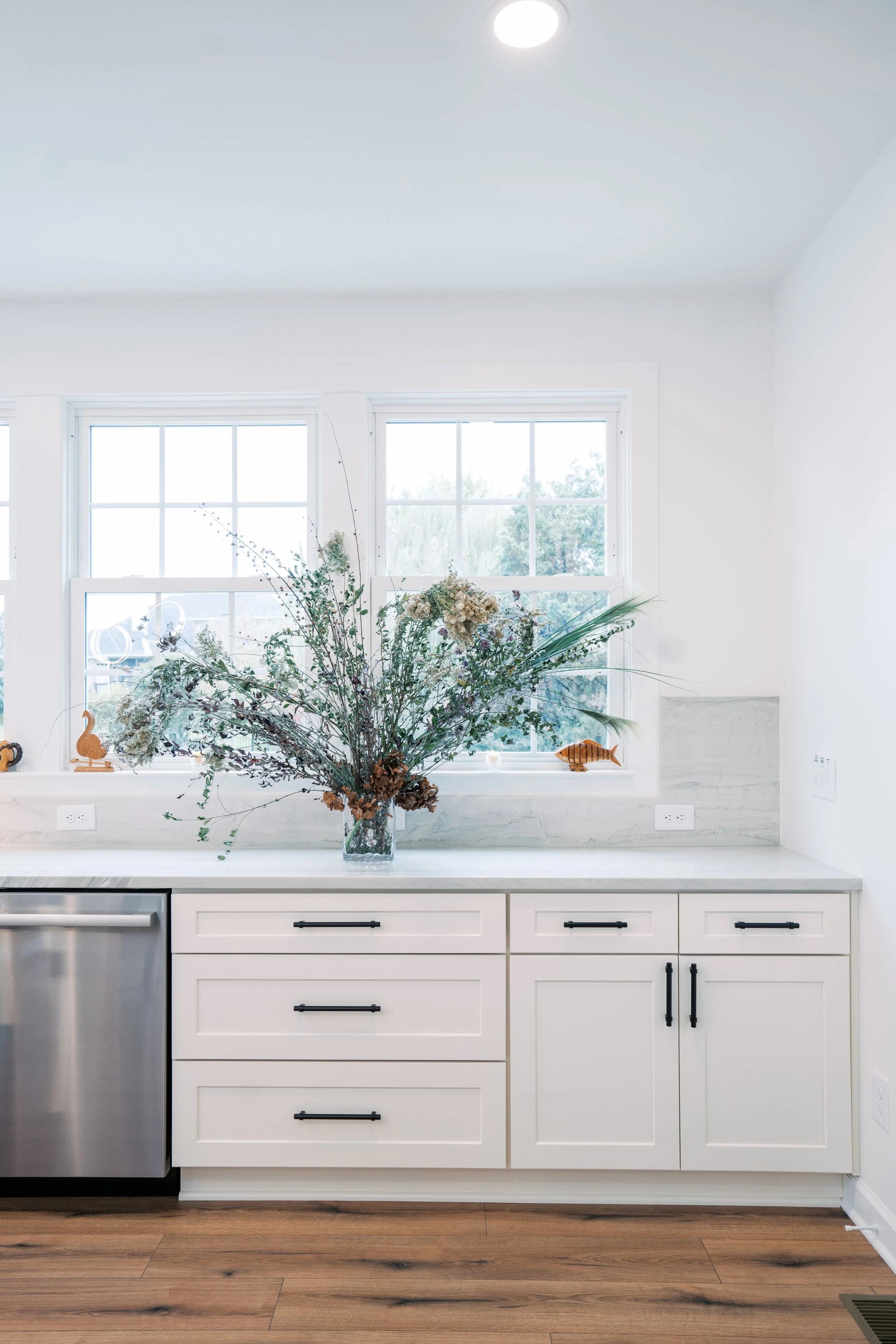 Kitchen countertop with a large vase of dried flowers, two windows in the background, and decorative wood carvings of a duck and a fish on the windowsill.