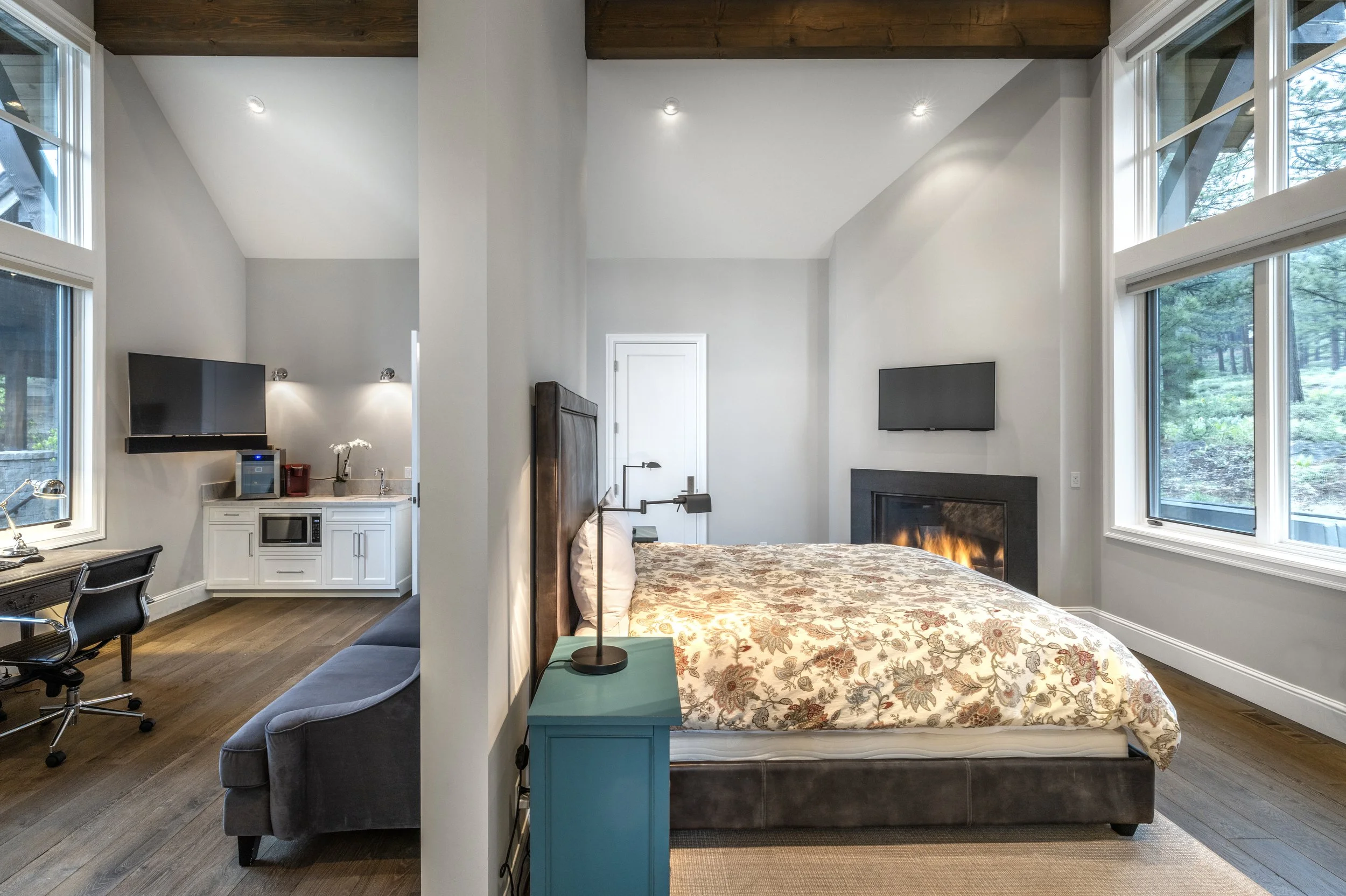 Interior of a modern bedroom with a large bed, nightstand, fireplace, flat-screen TV, and windows showing a wooded outdoor view.