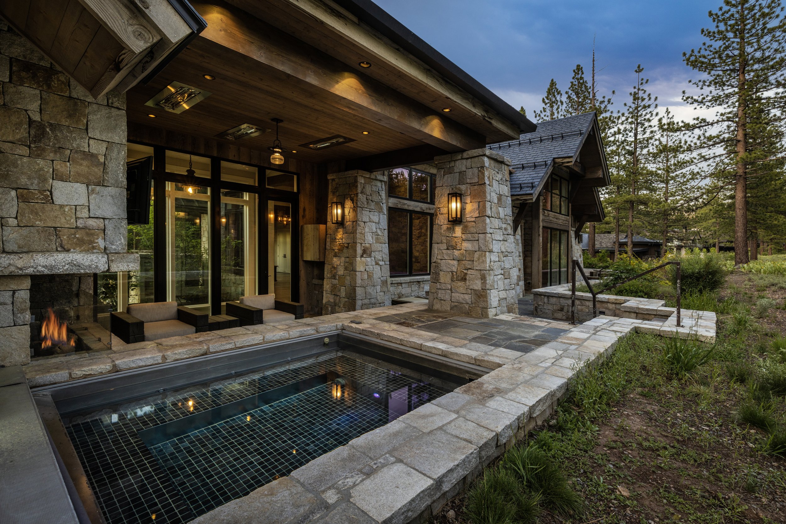 Exterior view of a modern stone and wood house with a small swimming pool, outdoor seating, and a fire pit, surrounded by trees during dusk.