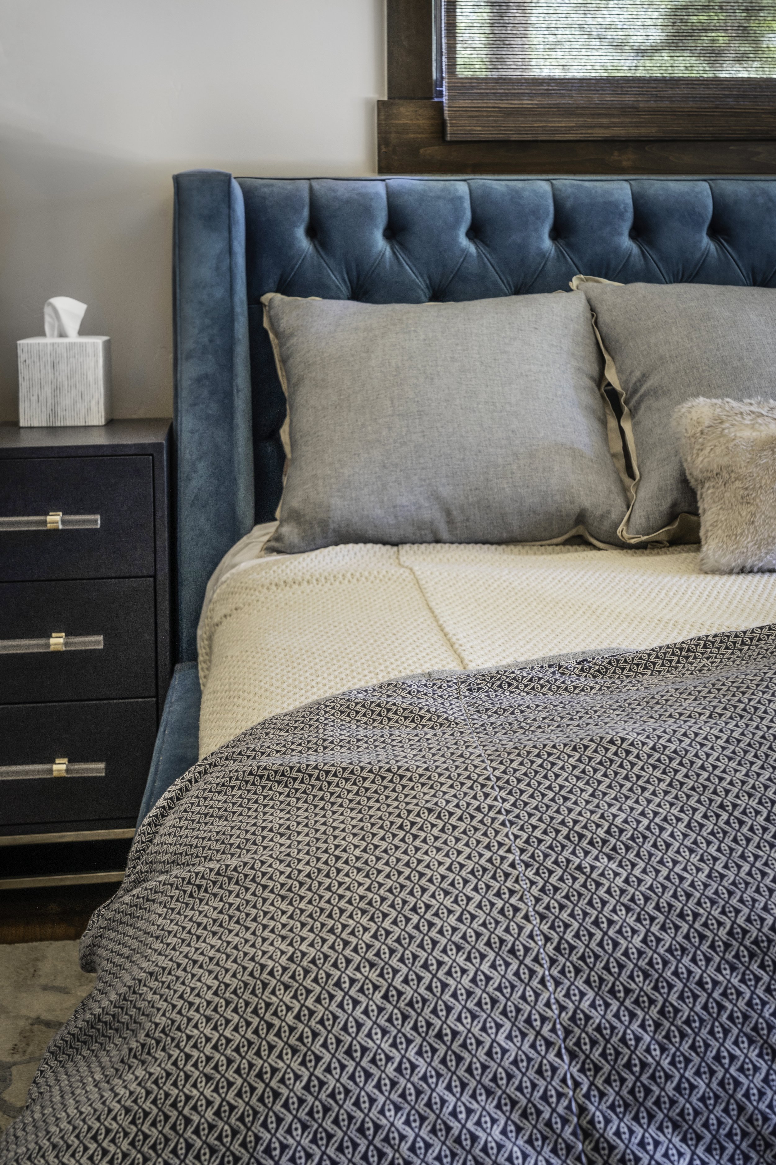 A bedroom with a teal upholstered headboard, gray pillows, and patterned bedding. Nightstand with a tissue box and a window with wooden blinds.
