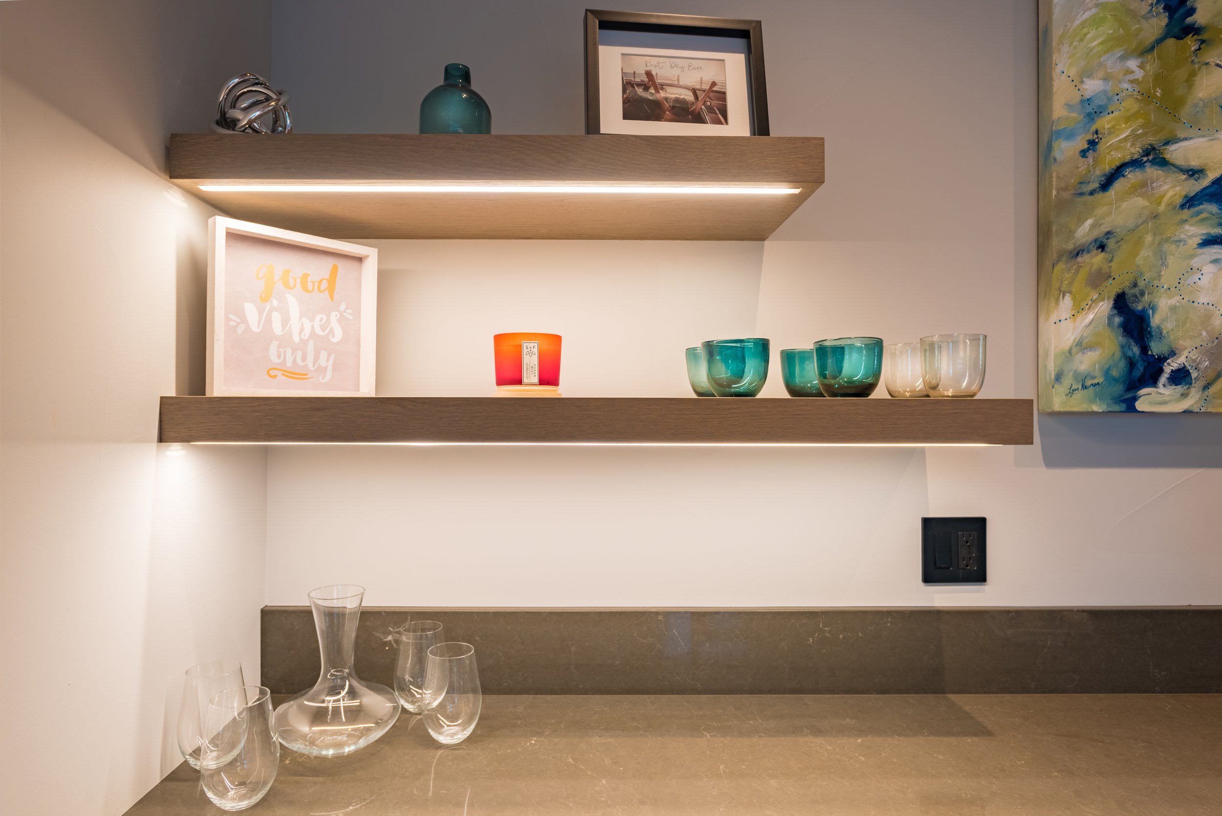Two floating wooden shelves with decorative items, including a framed quote, a framed photograph, a teal vase, a colorful candle, and glassware, against a wall with abstract art.