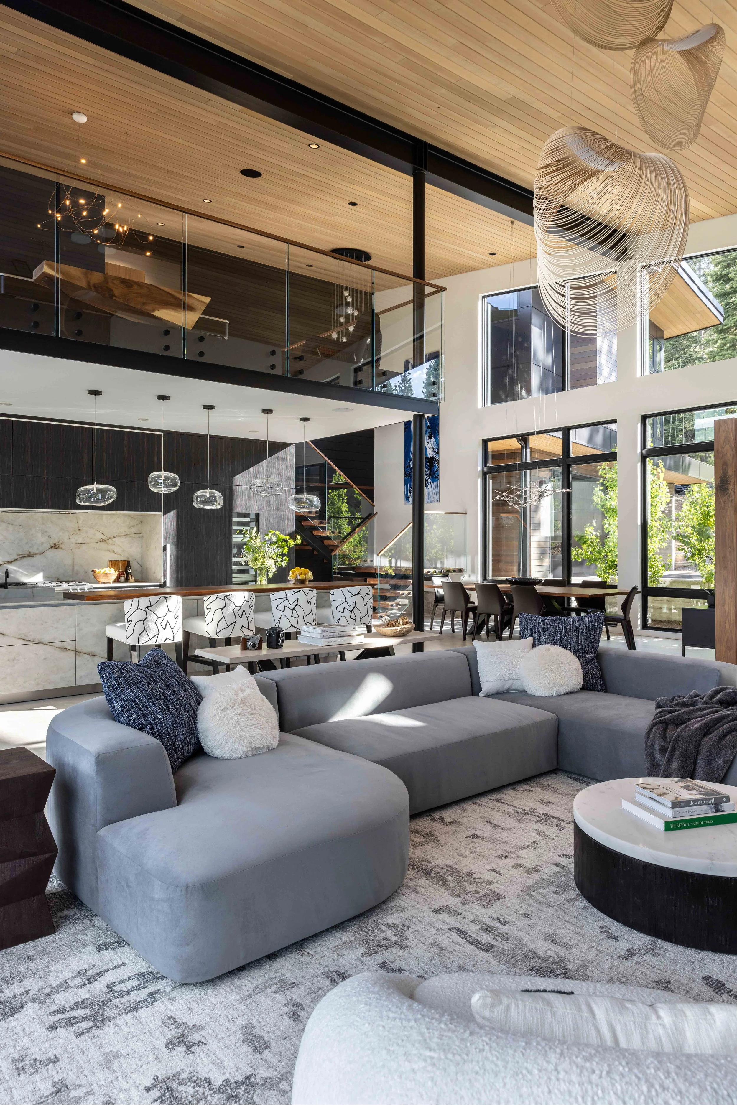 Open concept modern living room with high ceilings, large windows, gray sectional sofa with white and dark pillows, round marble coffee table with books, chandelier, and wood accents.