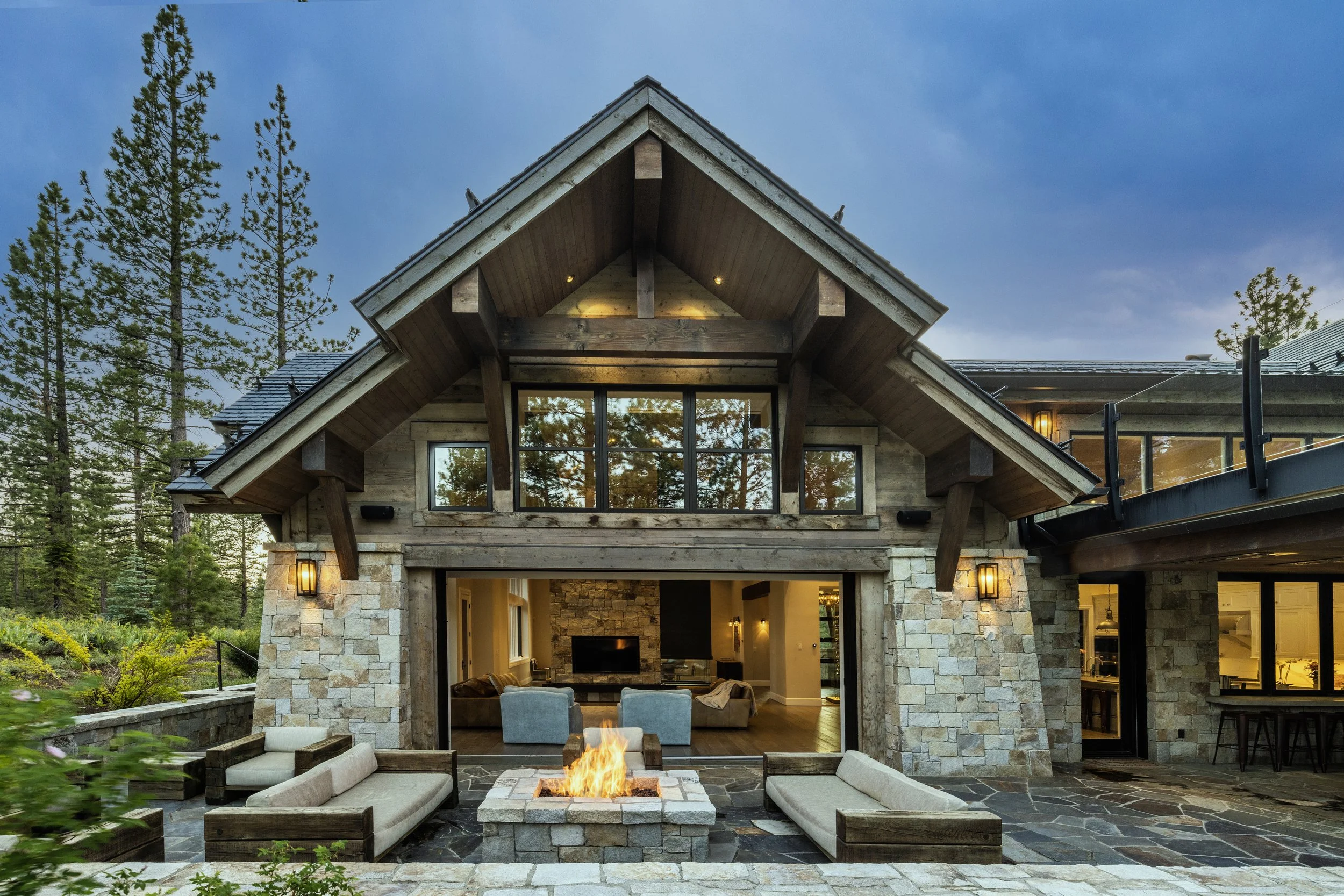 Luxury mountain home with stone and wood exterior, large windows, outdoor fireplace, and patio with seating, surrounded by tall pine trees at dusk.