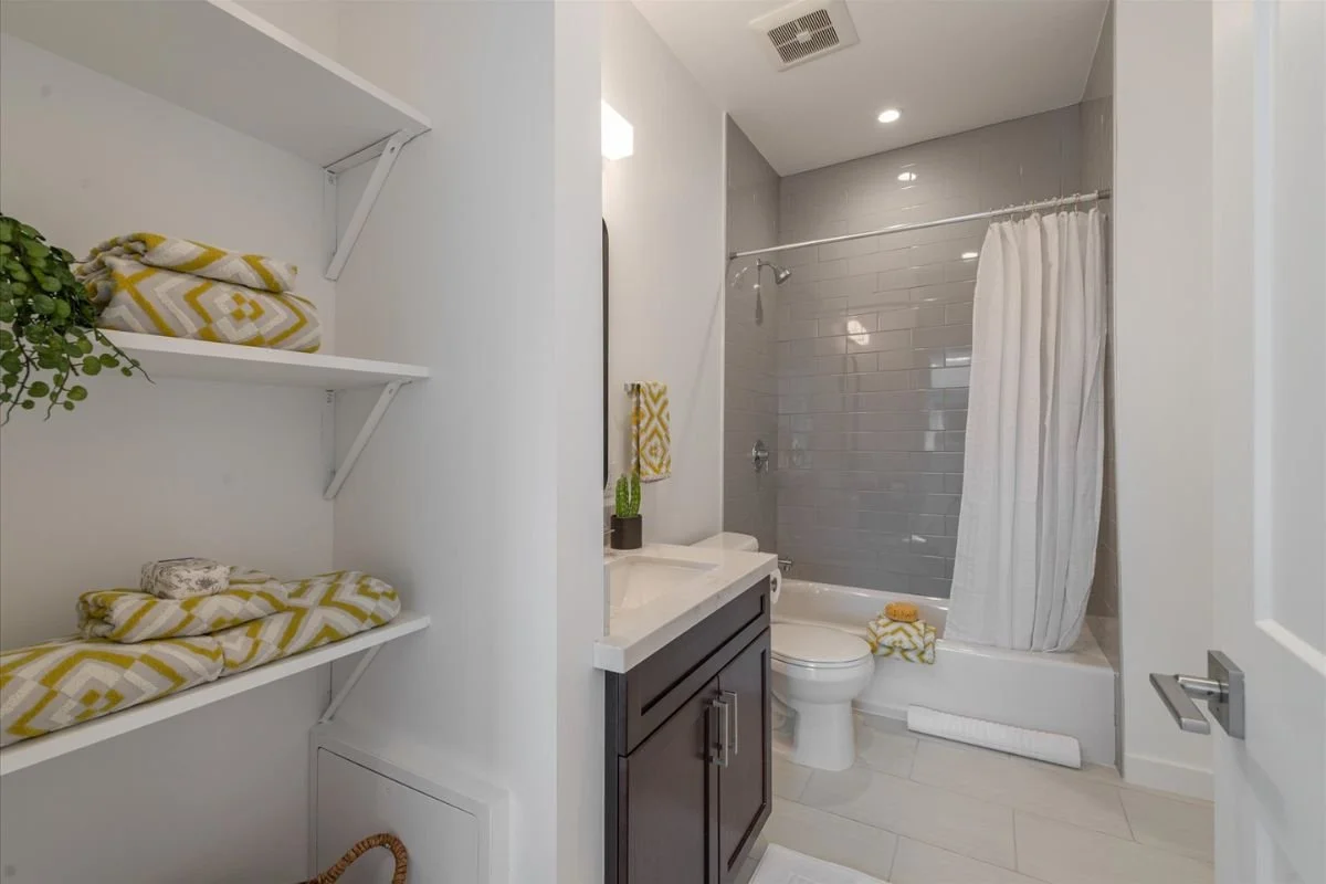 A modern bathroom with a shower area, a vanity with a sink, a mirror, and shelves with neatly folded towels and a plant.
