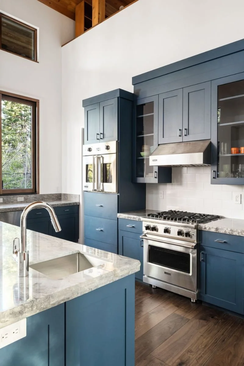 Modern kitchen with blue cabinets, stainless steel oven and range, marble countertops, and a window showing trees outside.