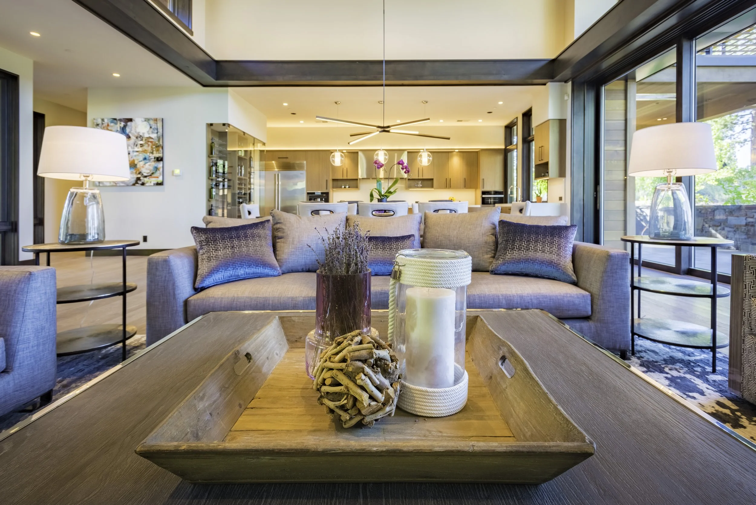 Living room with a gray sofa, wooden coffee table with decorative candles and dried plant, side tables with lamps, open kitchen in the background with wooden cabinets, large windows bringing in natural light.