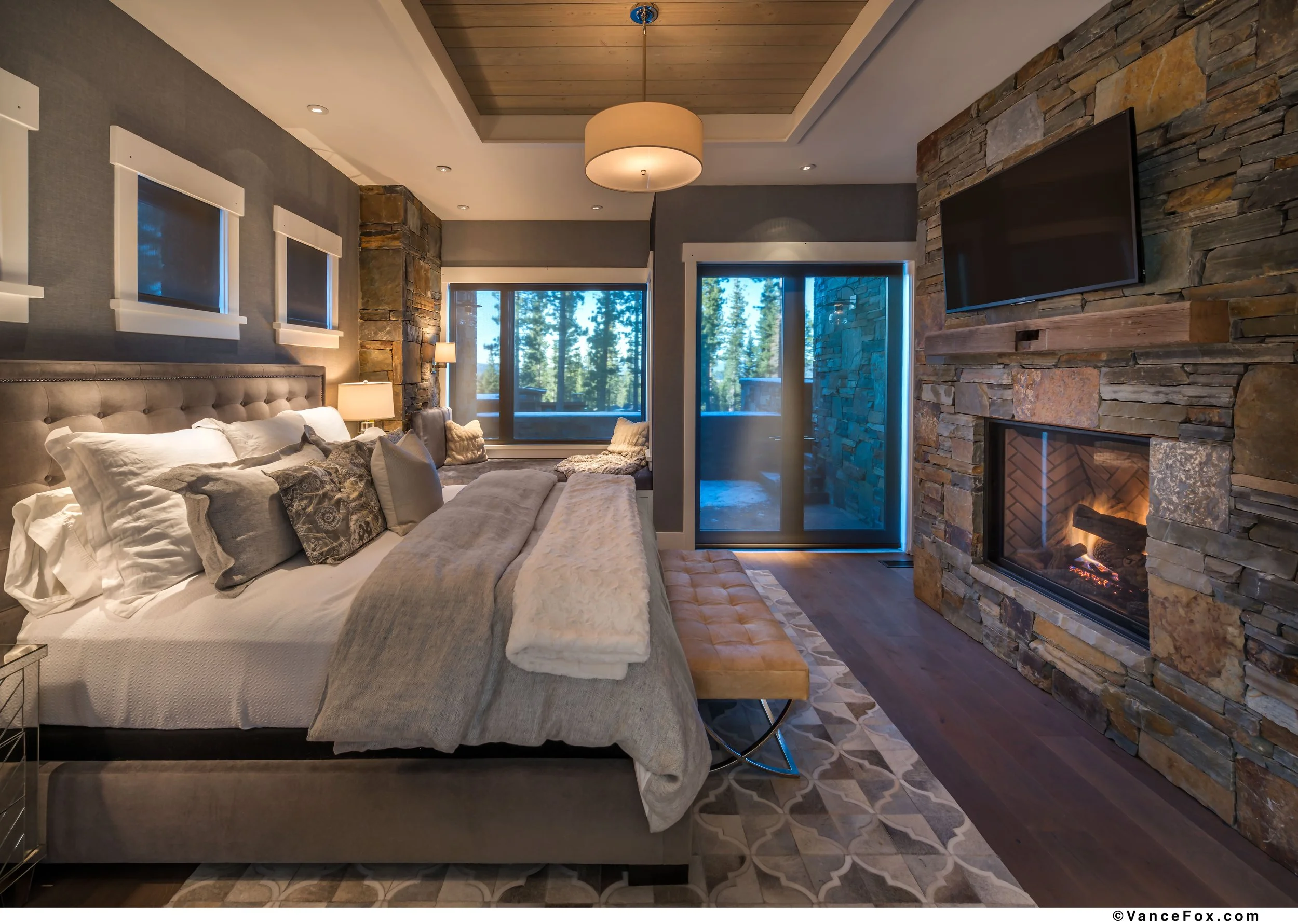Cozy bedroom with a large bed, gray upholstered headboard, multiple pillows, gray blanket, and faux fur throw. There are three small windows with blinds, a sliding glass door leading to a balcony, and a stone fireplace with a mounted flat-screen TV a