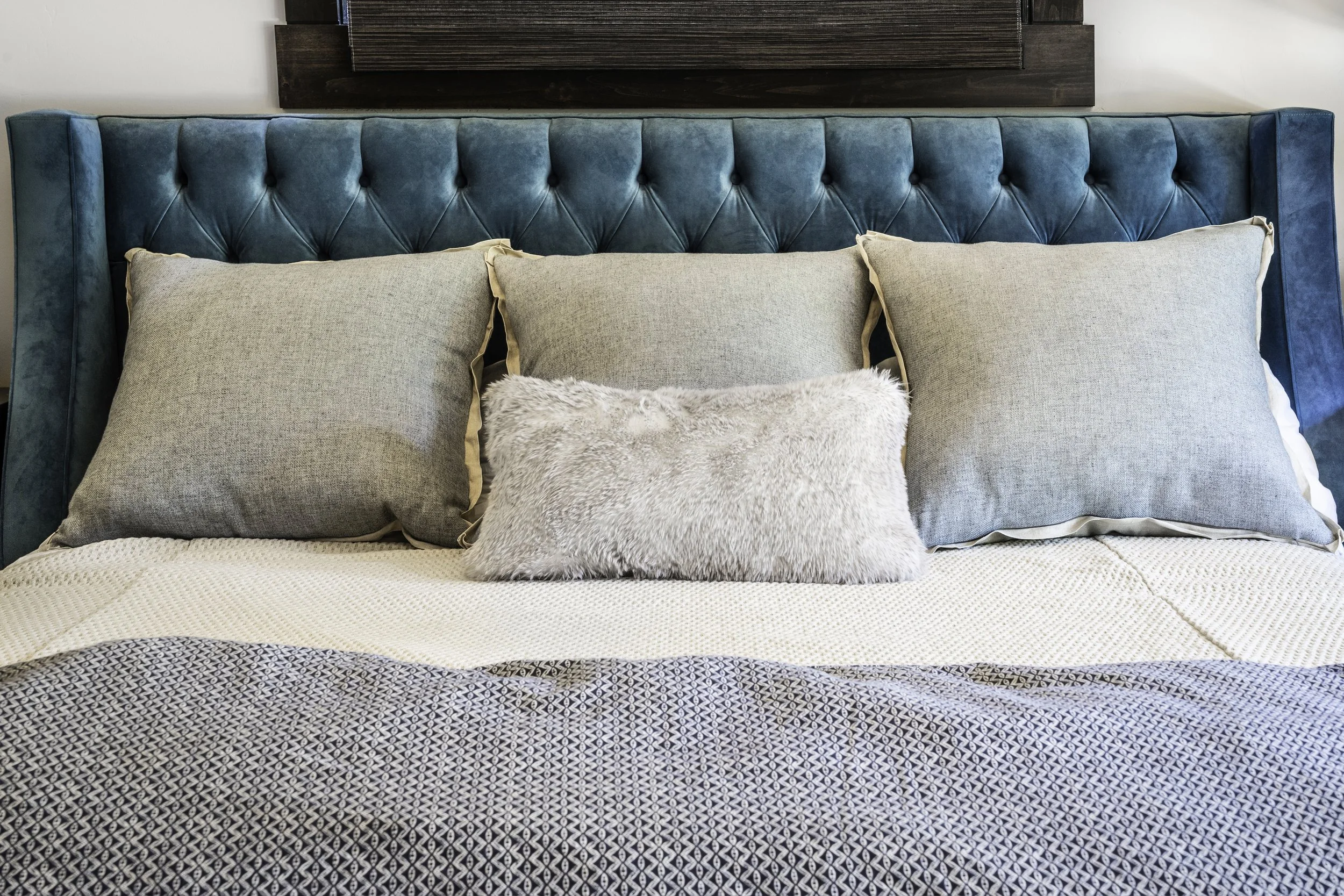 A bed with a turquoise upholstered headboard, three beige pillows with satin edges, and a gray furry bolster pillow. The bed has a gray and white patterned blanket.