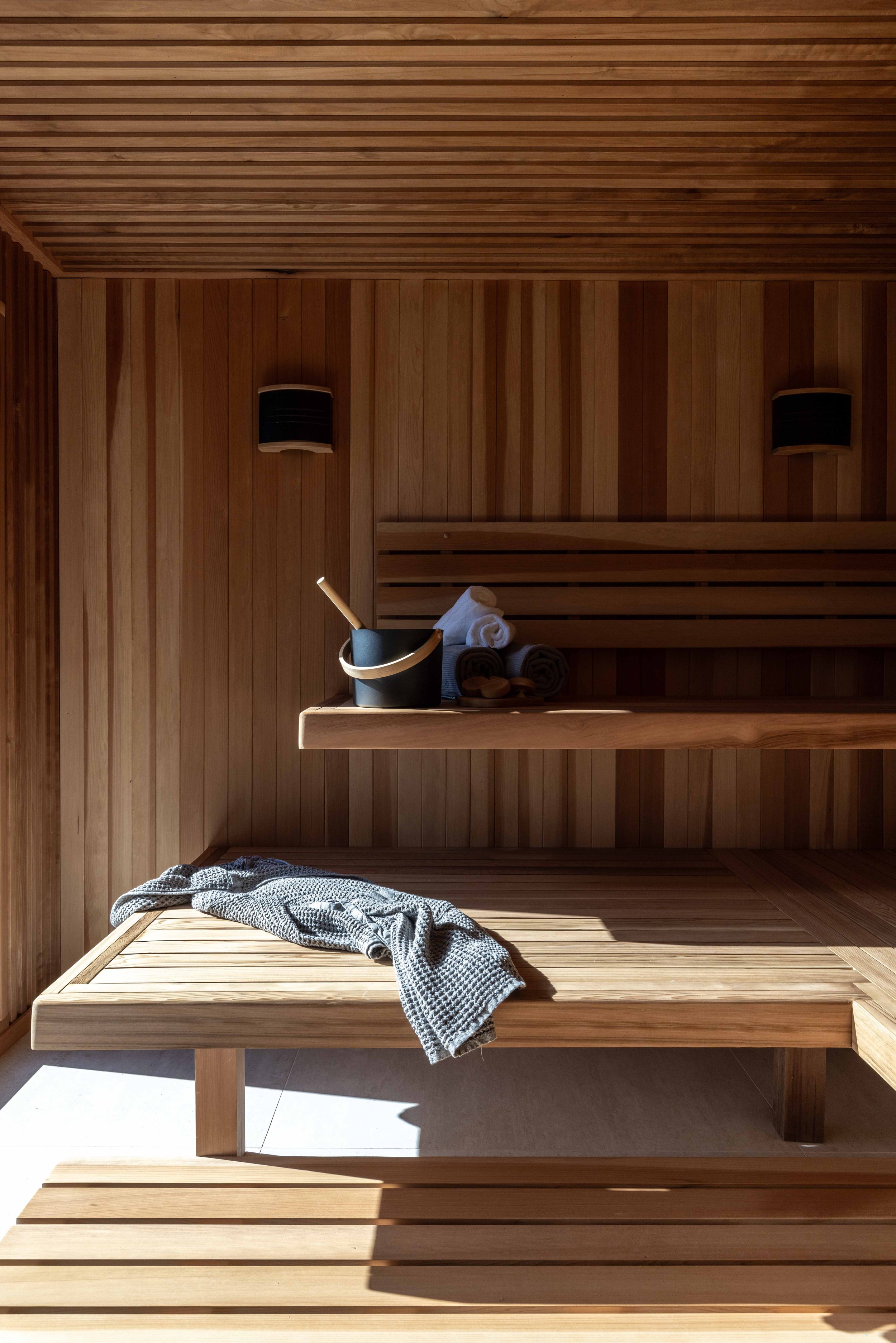 Interior of a wooden sauna with a bench, towels, a bucket with a ladle, and a gray towel draped over the bench.