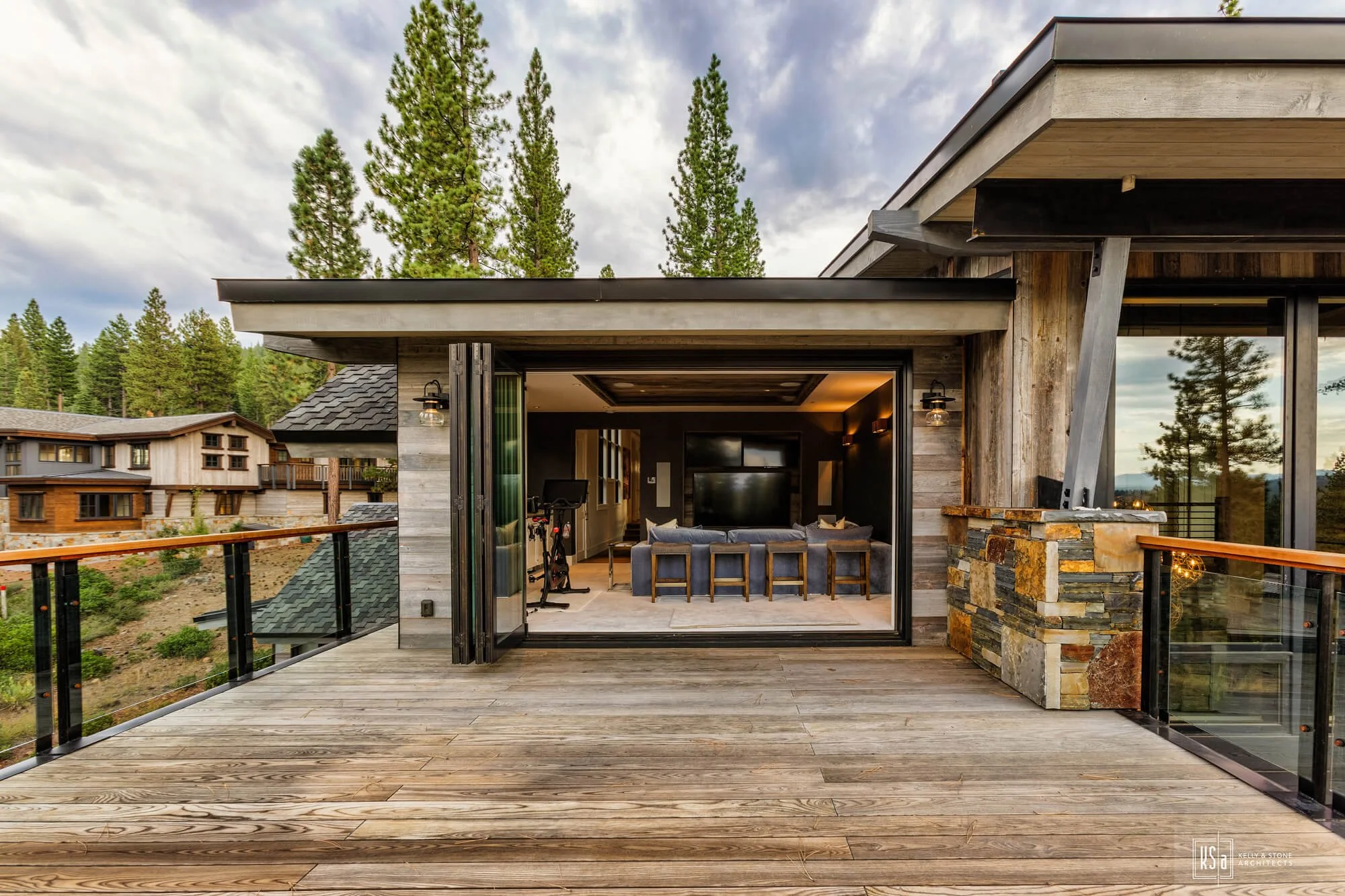 View of a modern house with a spacious wooden balcony opening to a living room with a large TV and seating area, surrounded by trees and scenic mountains.