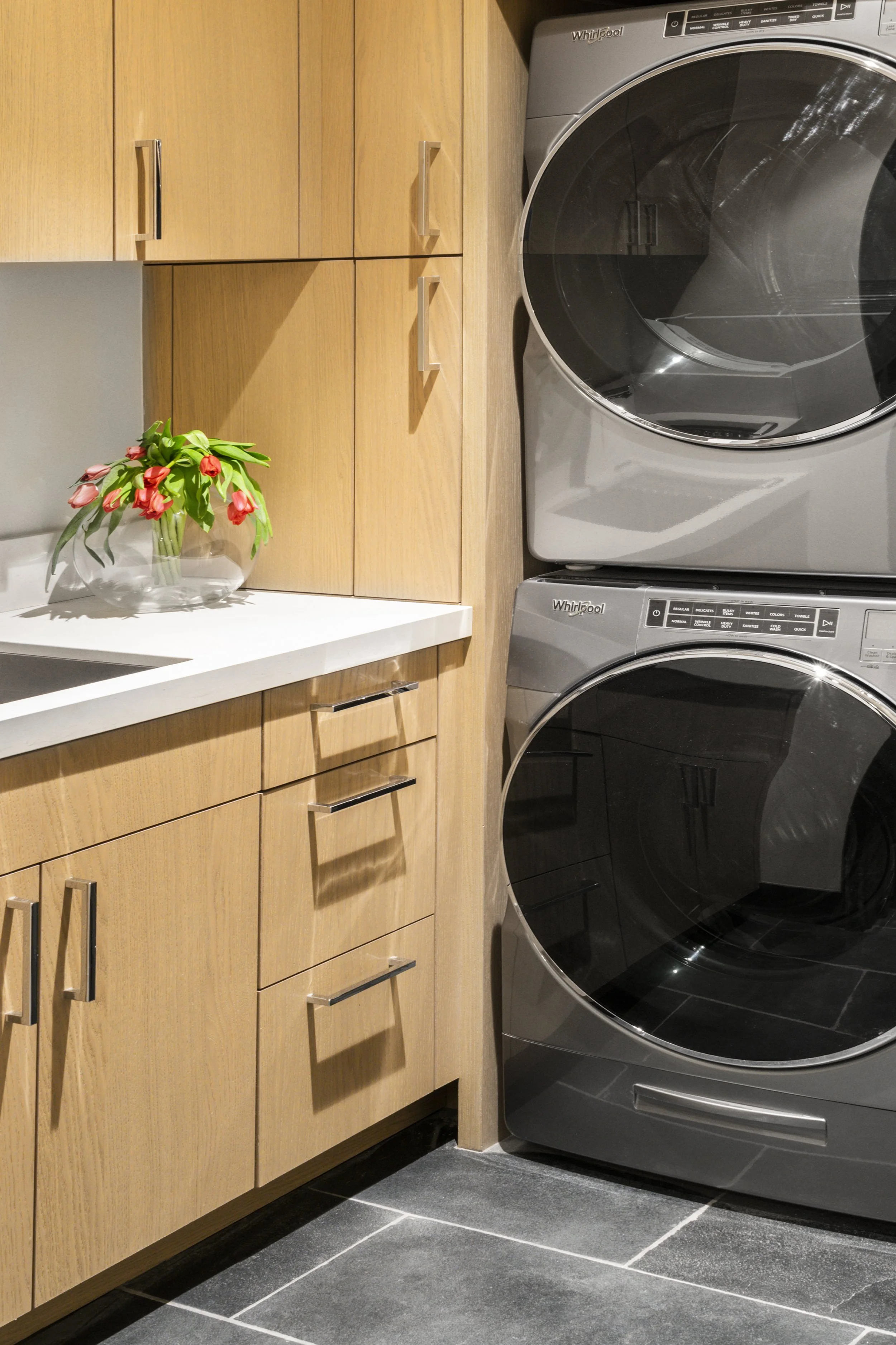A laundry room with wooden cabinets, a white countertop, a vase with red tulips, and a stacked washer and dryer set.