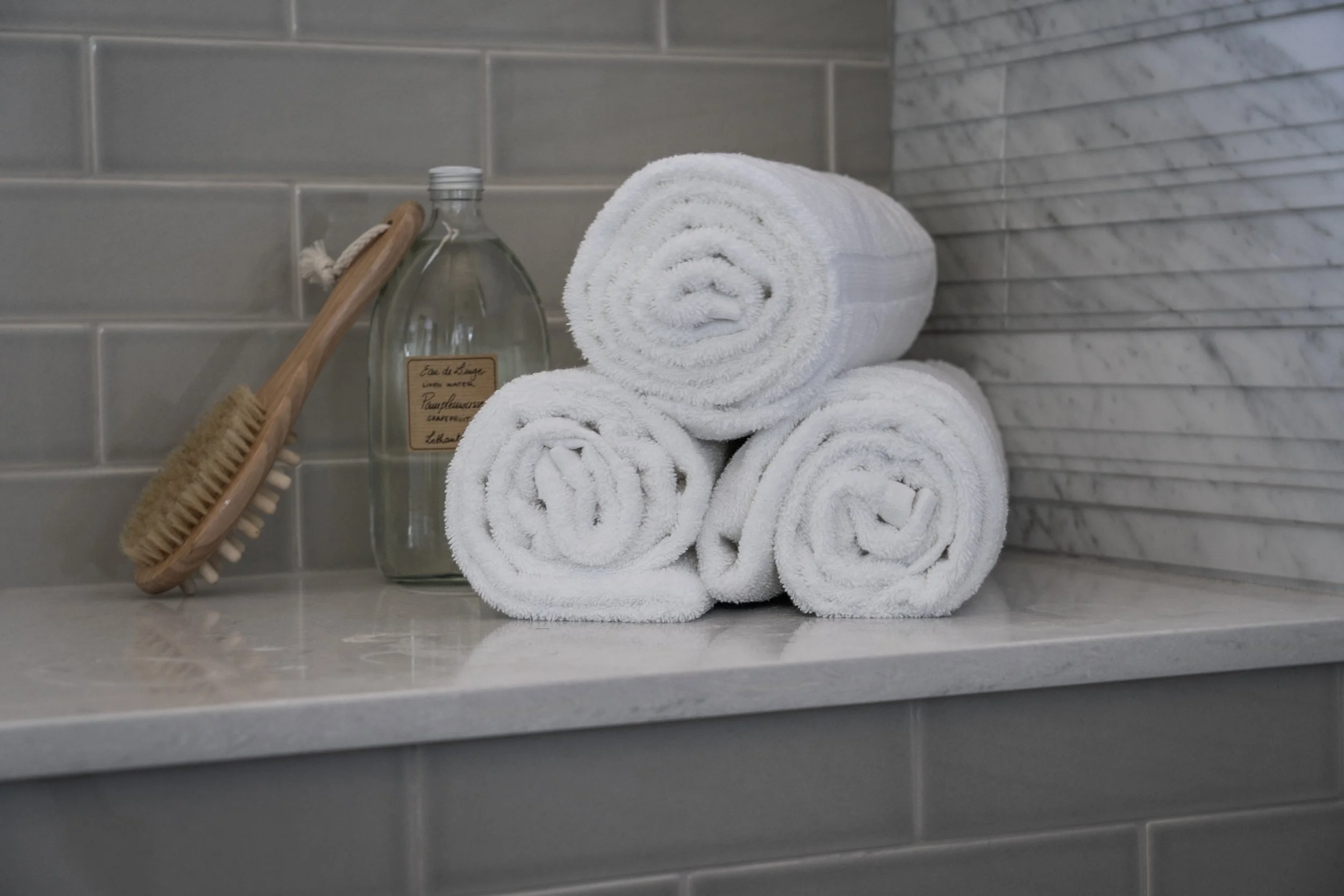 Stack of three folded white towels on a bathroom countertop with a wooden bath brush and a clear glass bottle.