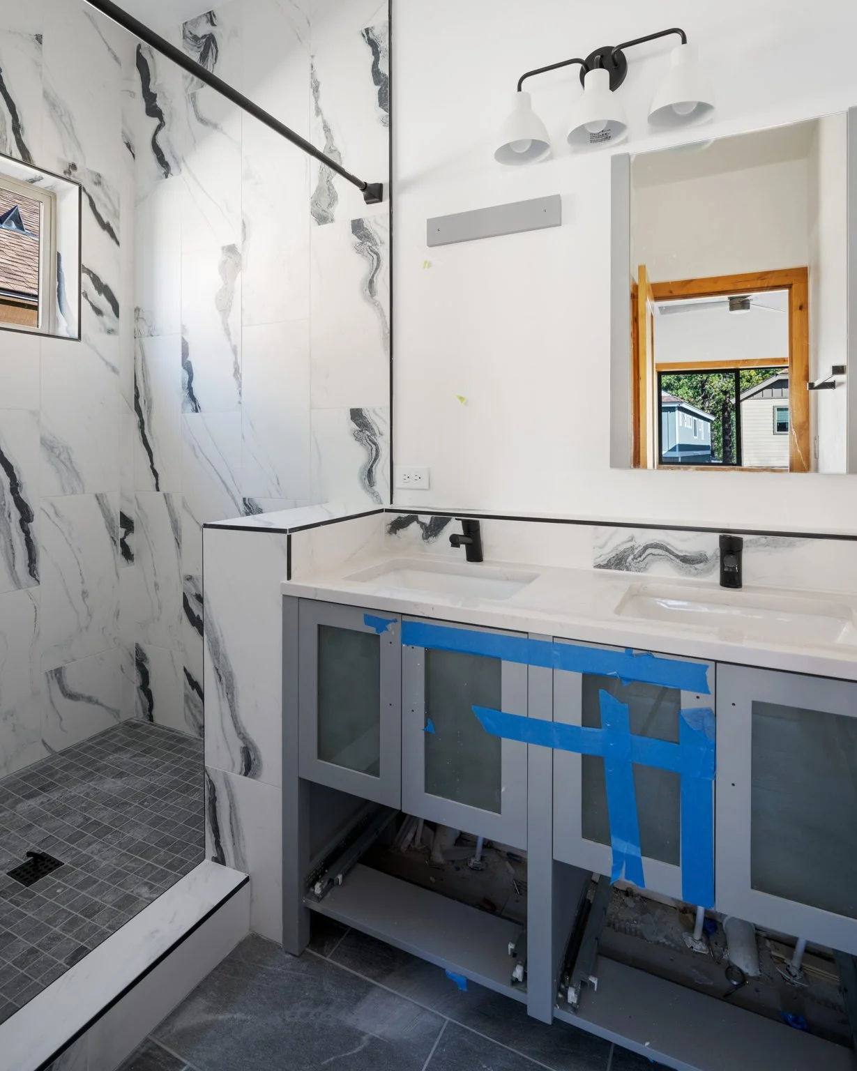 A bathroom under renovation with a partially installed double vanity cabinet, a marble tile shower, a wall mirror, and a window showing houses outside.