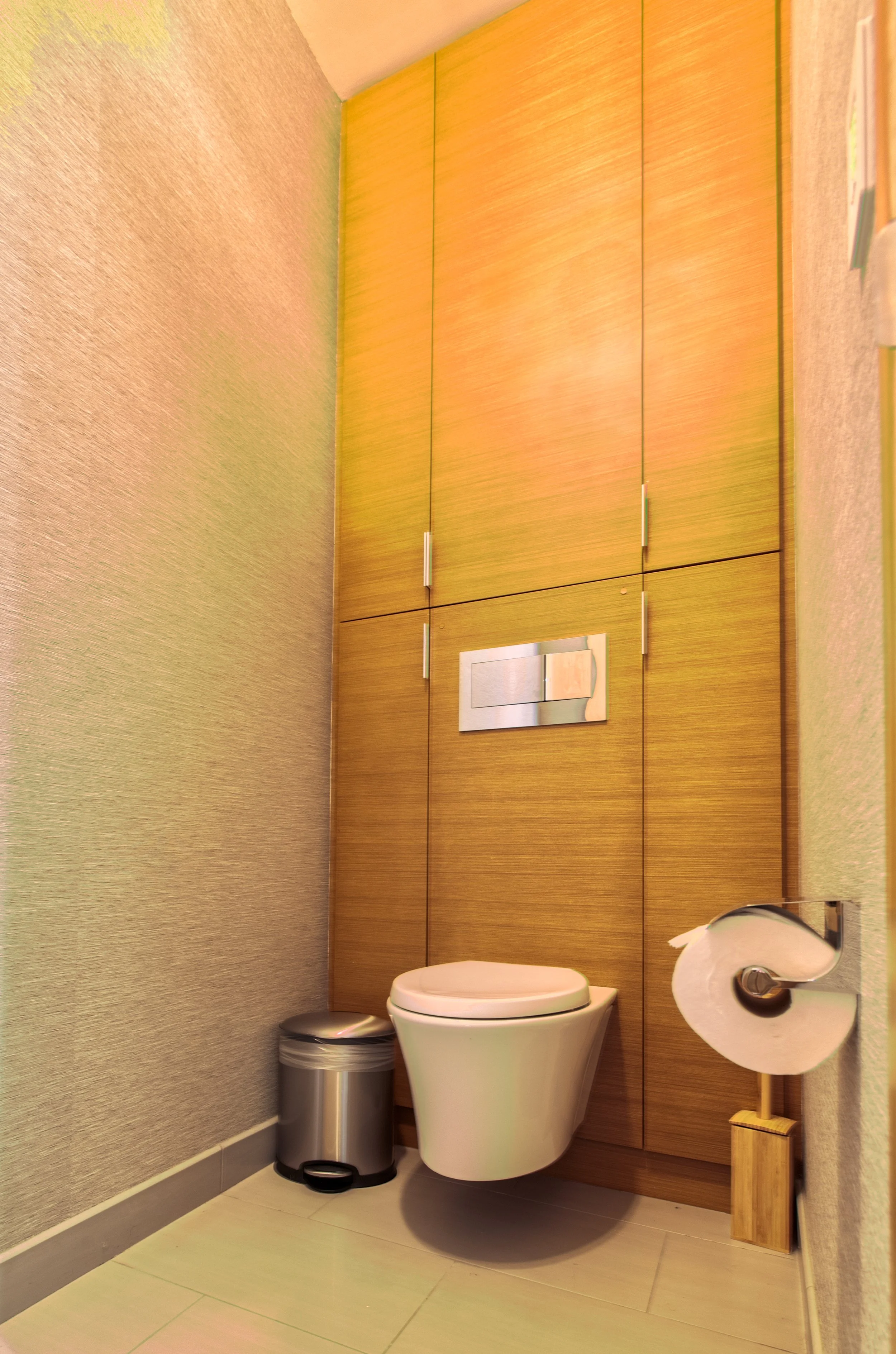 A modern bathroom with a wall-mounted toilet, a wooden cabinetry wall, a stainless steel trash bin, and a toilet paper roll on a hold with a toilet brush in a wooden holder.