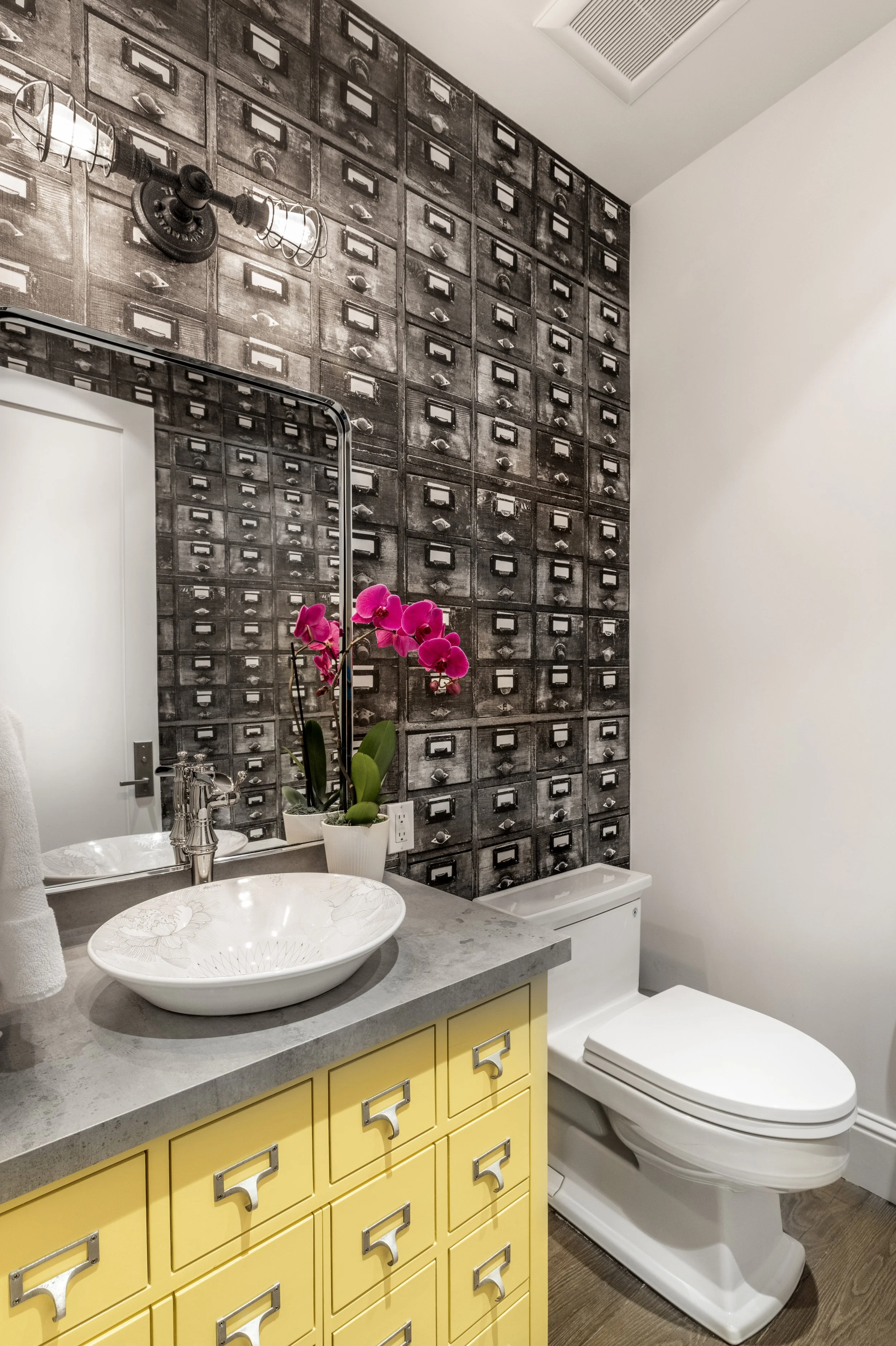 A modern bathroom with a yellow cabinet, a white vessel sink, a large mirror, and a black and white photo-inspired wall with multiple small drawers. There is a pink orchid plant on the counter, a towel hanging on the side, and a white toilet next to 