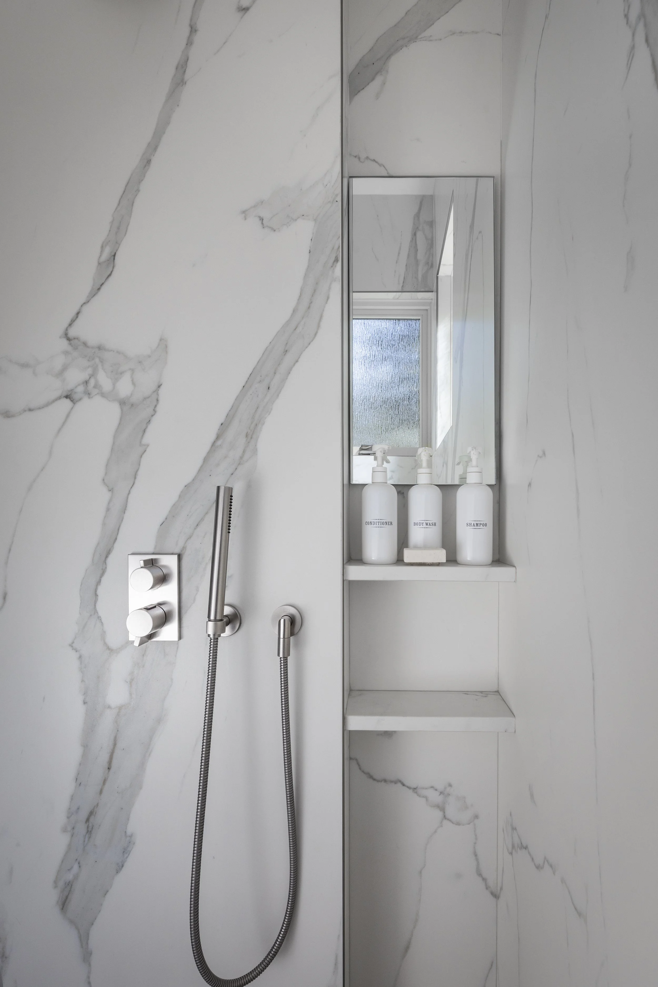 The primary shower walls are covered in large porcelain slabs to reduce grout lines and easy to clean as well as looking stunning and luxurious. An anti-fog mirror is added for ease of shaving with floating shelves for all shower needs. A handshower 