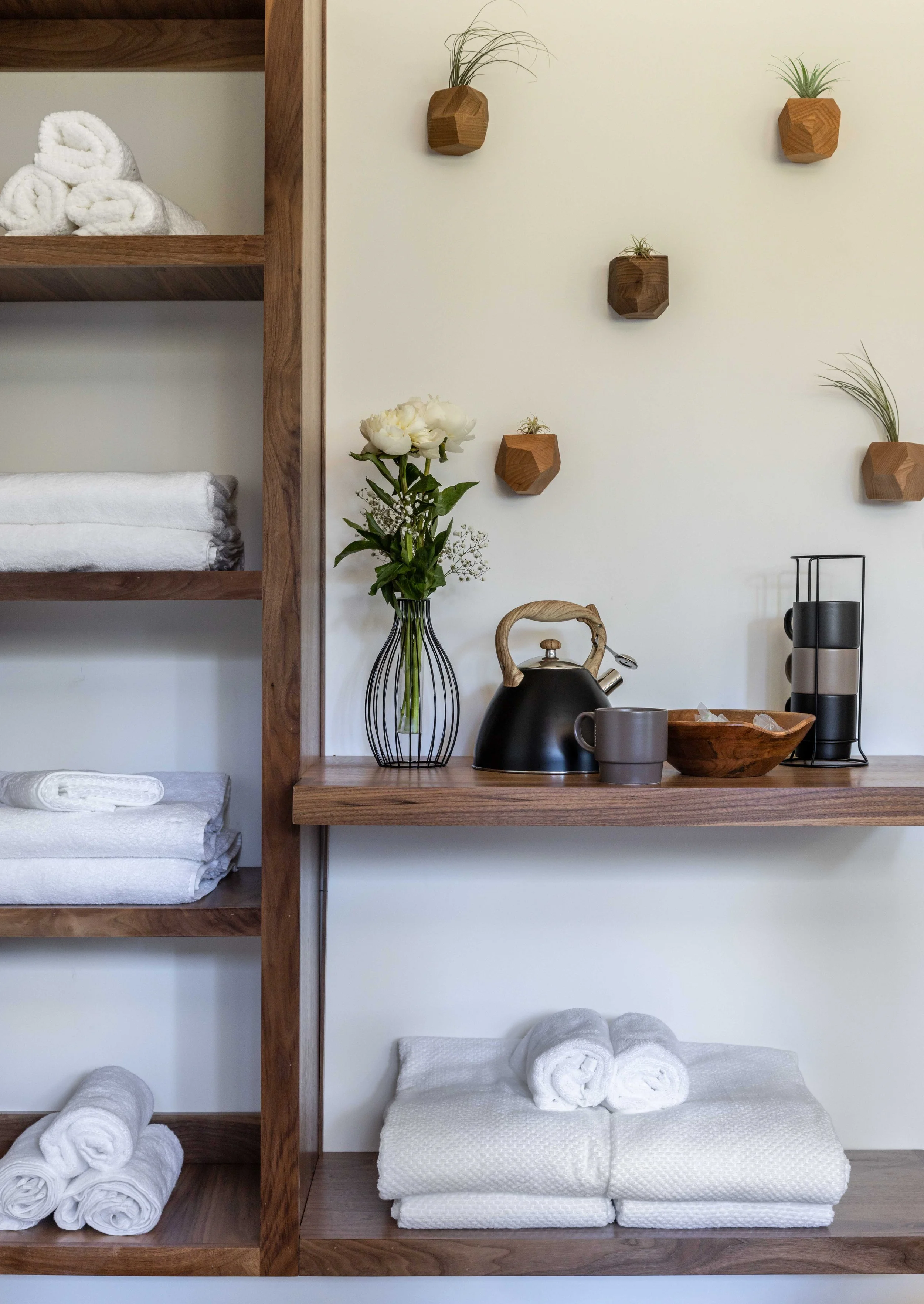 A wooden shelf with rolled white towels, a vase with white flowers, a black teapot, a gray mug, and a wooden bowl, against a white wall decorated with small planters with green plants.