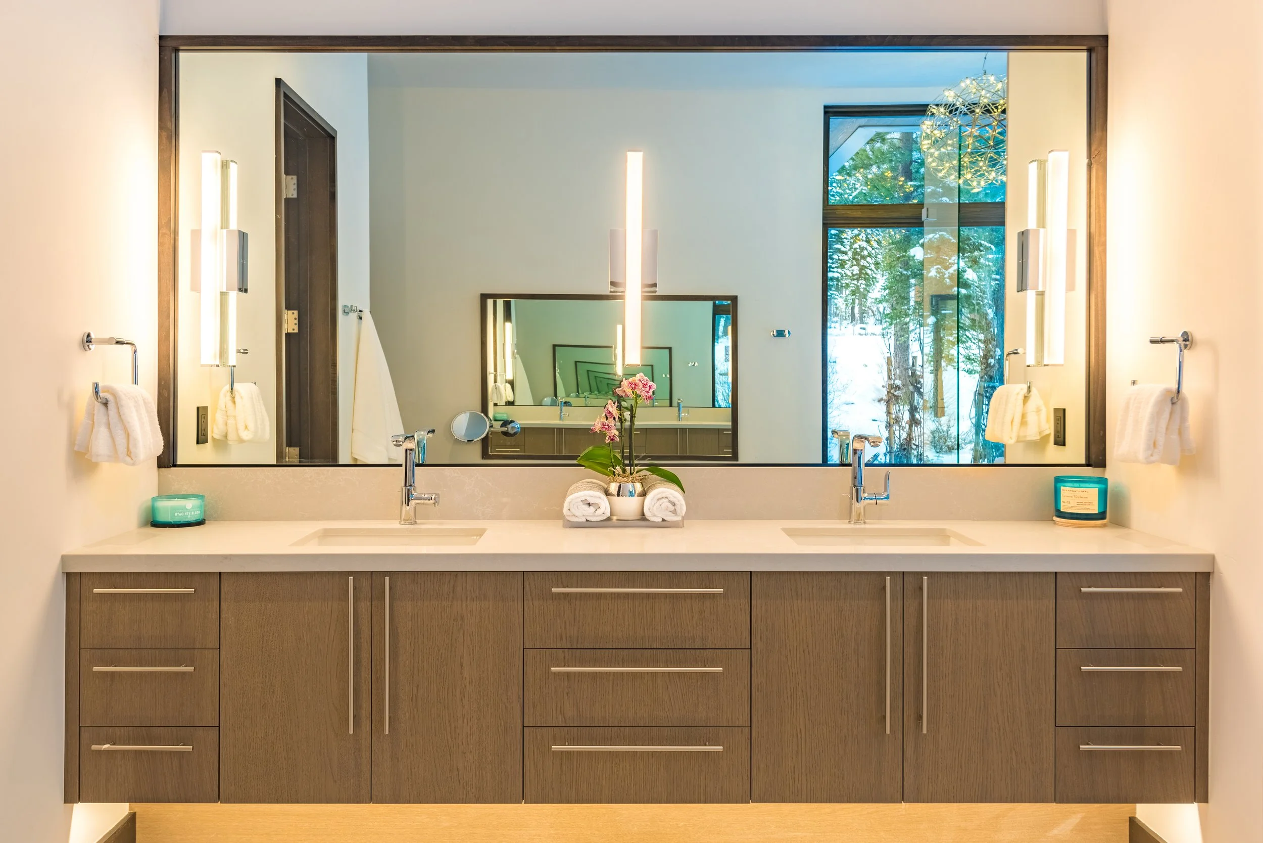Modern bathroom with double sinks, large mirror, wooden cabinets, decorated with a flower arrangement and rolled towels, illuminated by wall sconces, with windows showing greenery outside.