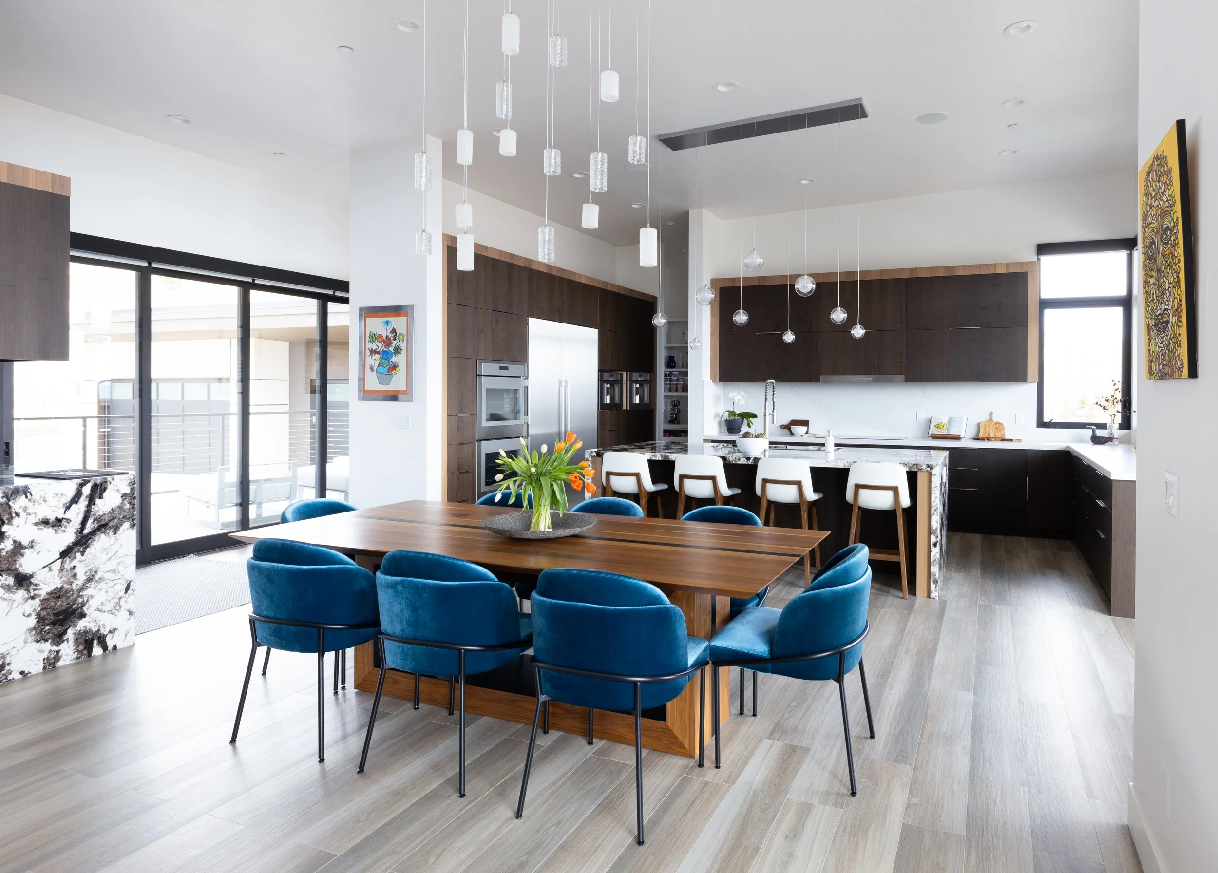 Modern kitchen and dining area with a wooden table and blue upholstered chairs, a kitchen island with white barstools, dark wood cabinetry, and large windows.