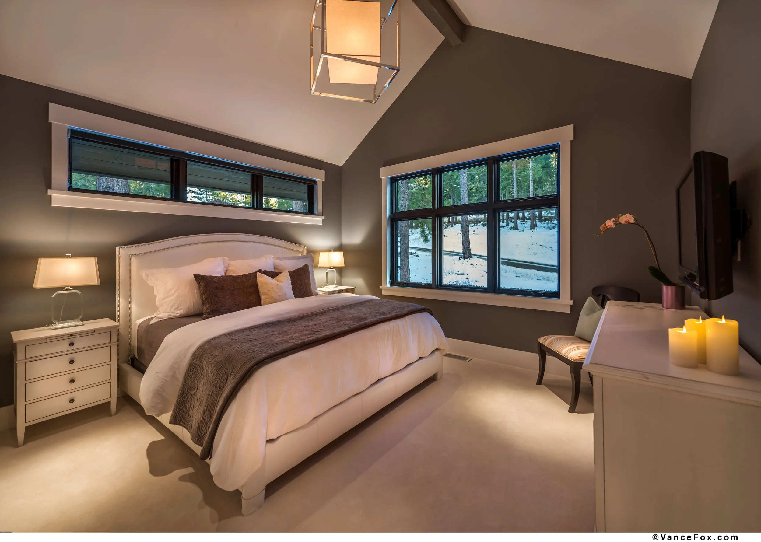 Cozy bedroom with a large bed, nightstands, lamps, and view of snow-covered trees outside through large windows.