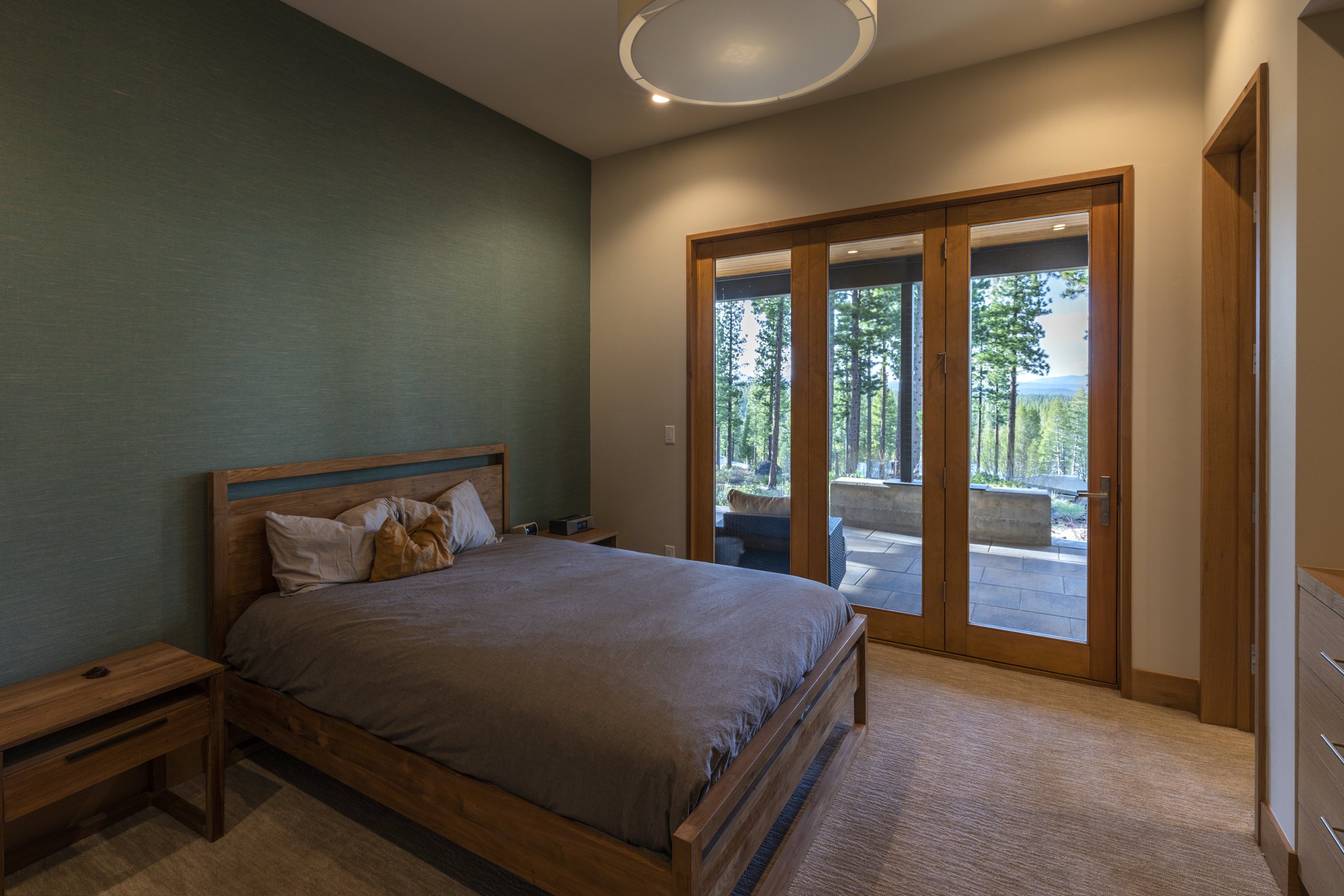 A bedroom with a wooden bed frame, brown bedding, and pillows, next to a wooden nightstand, large glass doors leading to a patio with outdoor seating, and a view of trees and mountains outside.