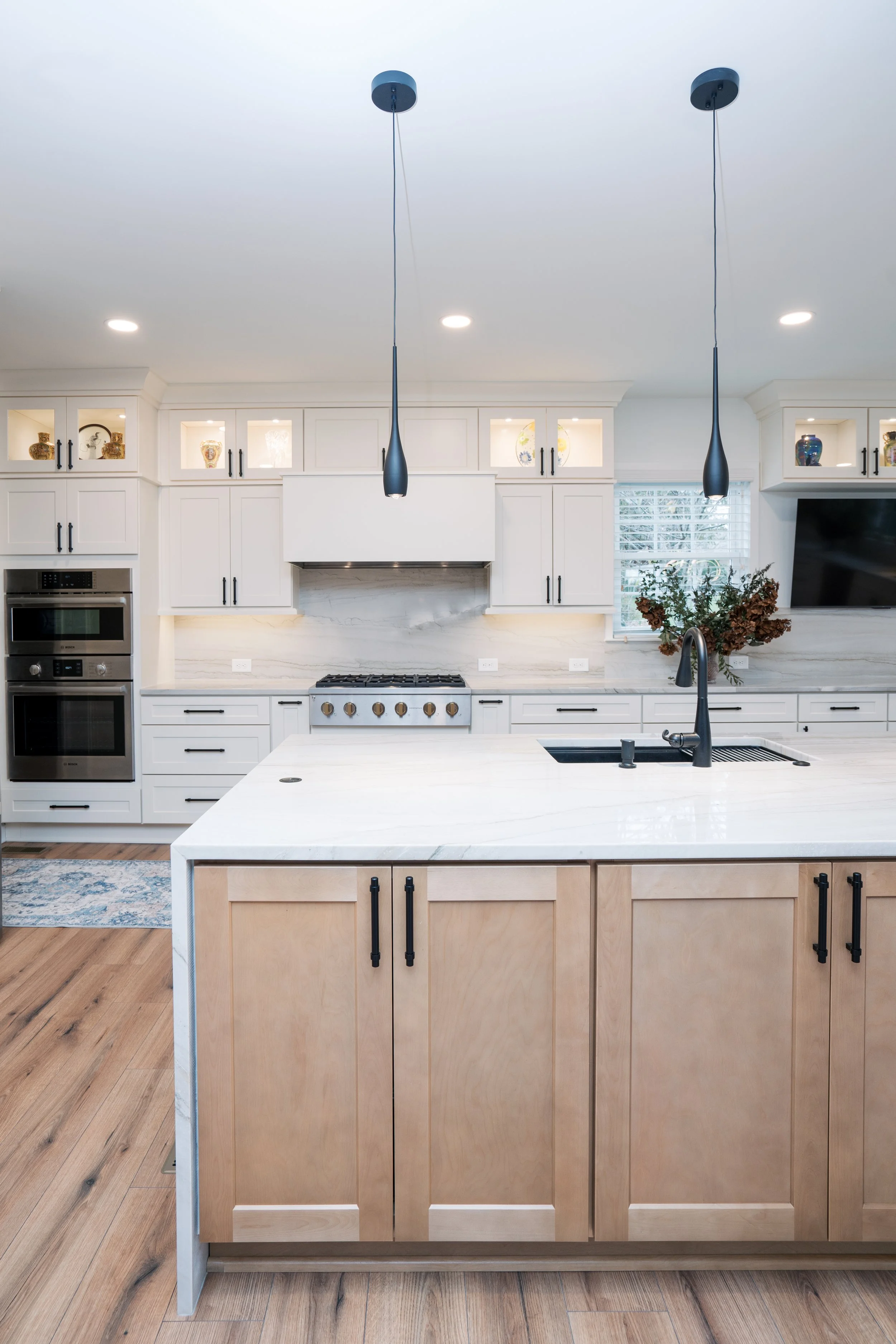 Transitional kitchen with white cabinets, a kitchen island with wooden cabinets, black fixtures including a faucet, hanging pendant lights, a stove, a microwave oven, a TV on the wall, and a vase with plants.
