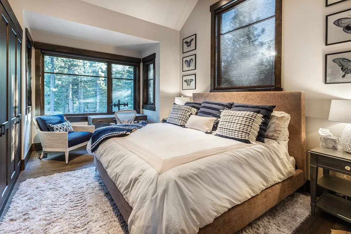 Cozy bedroom with large window, bed with beige headboard and pillows, side table with lamp, armchair with pillows, wall of framed butterfly art, soft rug.