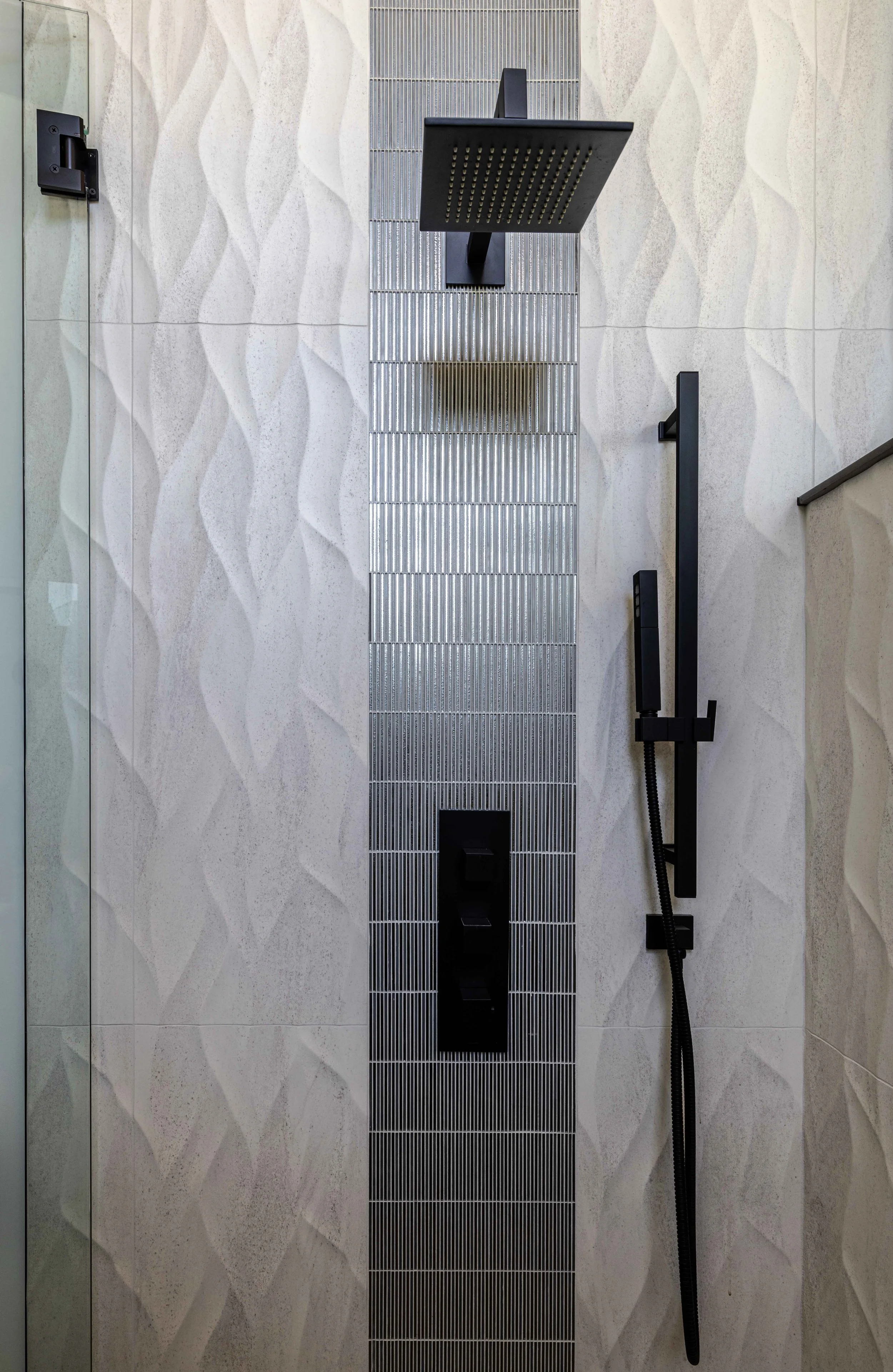 Shower with black square showerhead, handheld shower, and water control panel mounted on textured tiled wall.