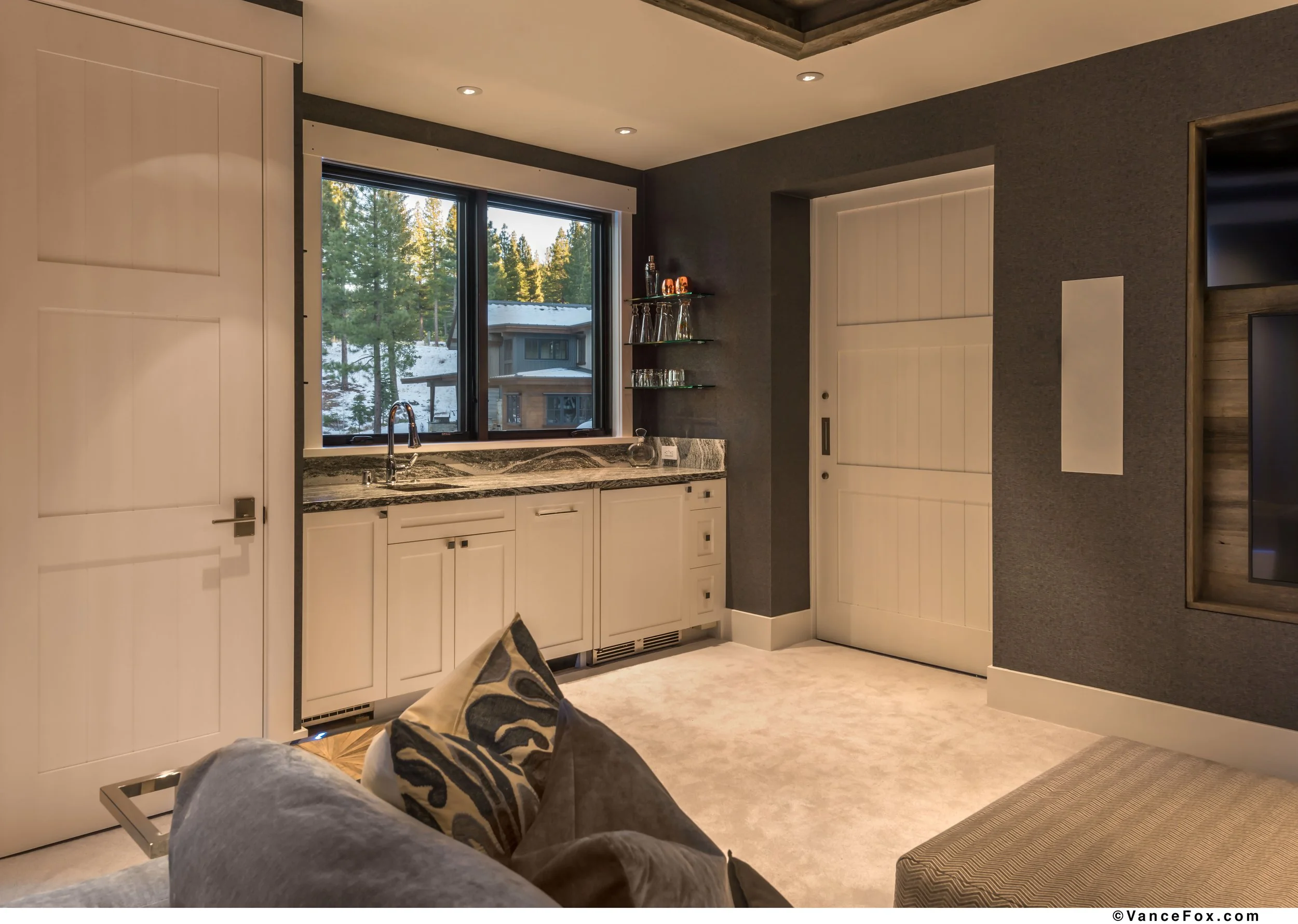 Kitchenette in a media room with a dishwasher, beverage fridge, and ice maker. The space is closed off by a sliding barn door, a wall-mounted televisions, and a denim sectional with patterned pillows.