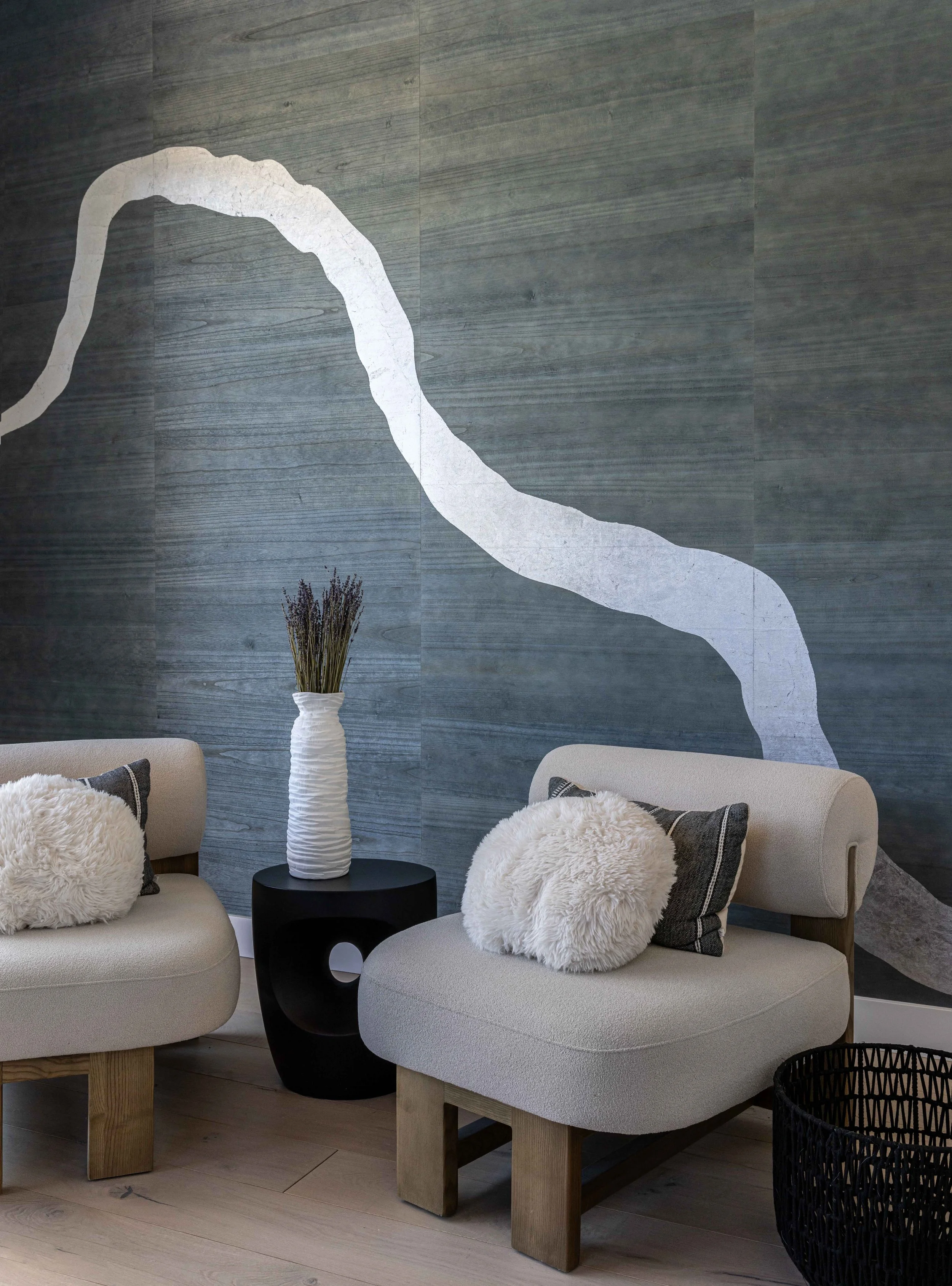 Modern living room with gray textured wall featuring a large white abstract stripe, two beige upholstered chairs with decorative pillows, a black side table with a white textured vase containing lavender, and a black woven basket.