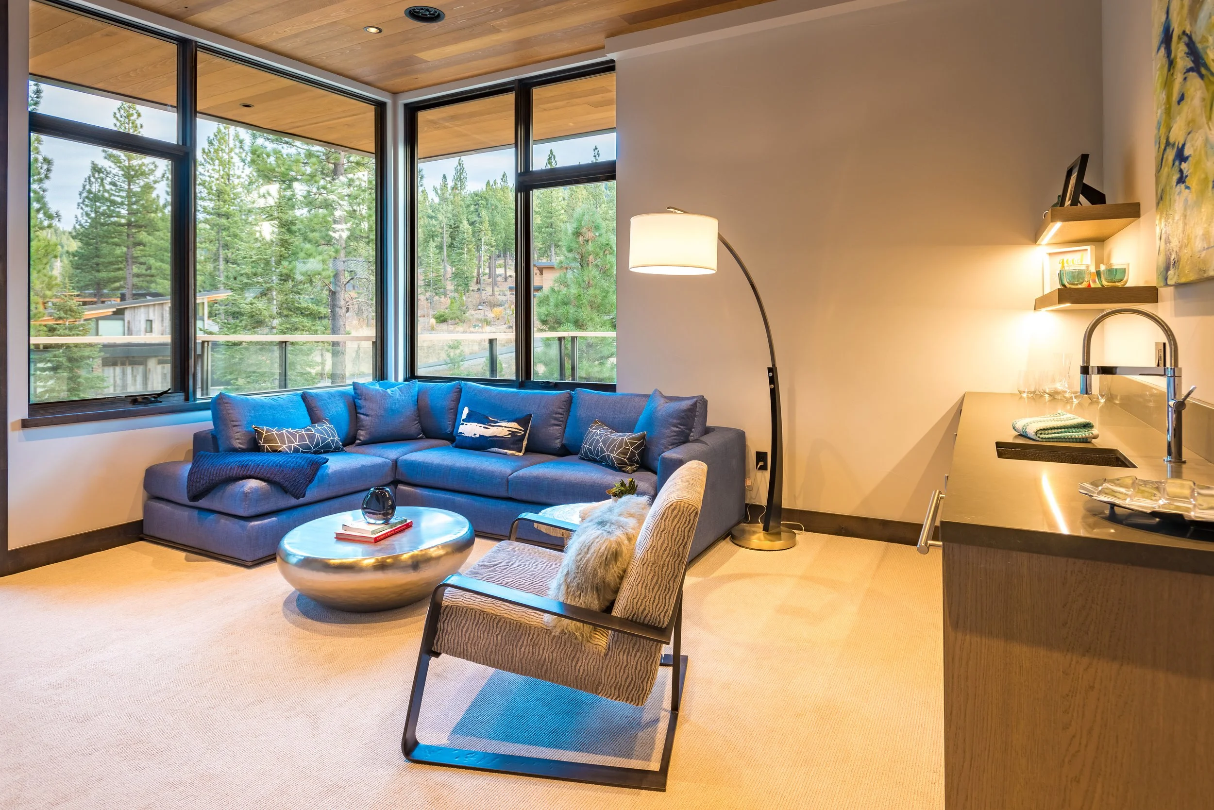 Modern living room with large windows overlooking a wooded landscape, blue sectional sofa with multiple cushions, beige armchair with fur throw, round metallic coffee table, beige carpet, and partial view of a kitchen counter with glasses and a towel