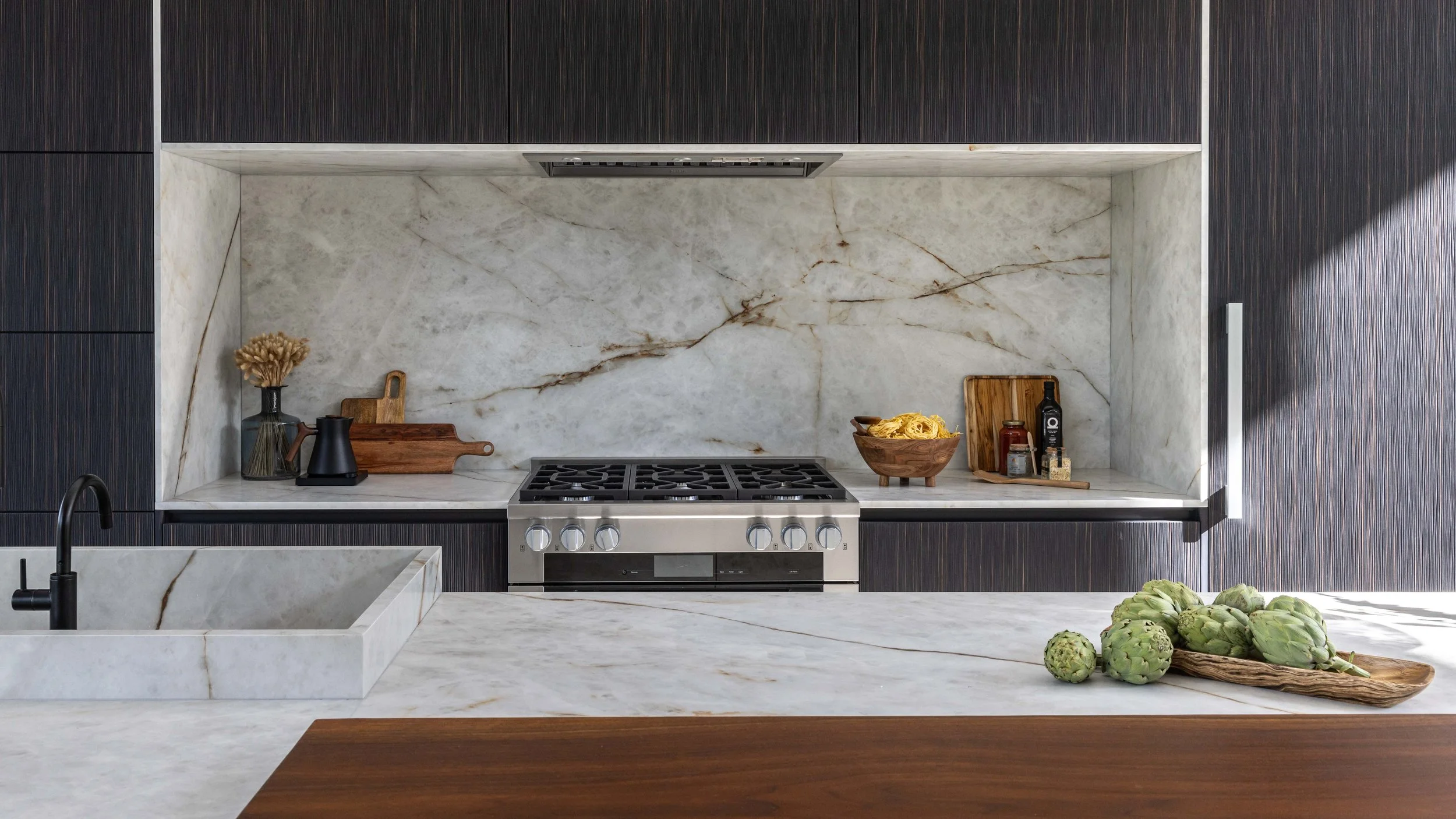 Organic Mountain Modern kitchen with Himalaya Crystal porcelain countertops and backsplash with a live edge walnut counter suspended on the island, black and brown cabinets mixed with dark brown accents, a Wolf range, and artichokes on the counter.