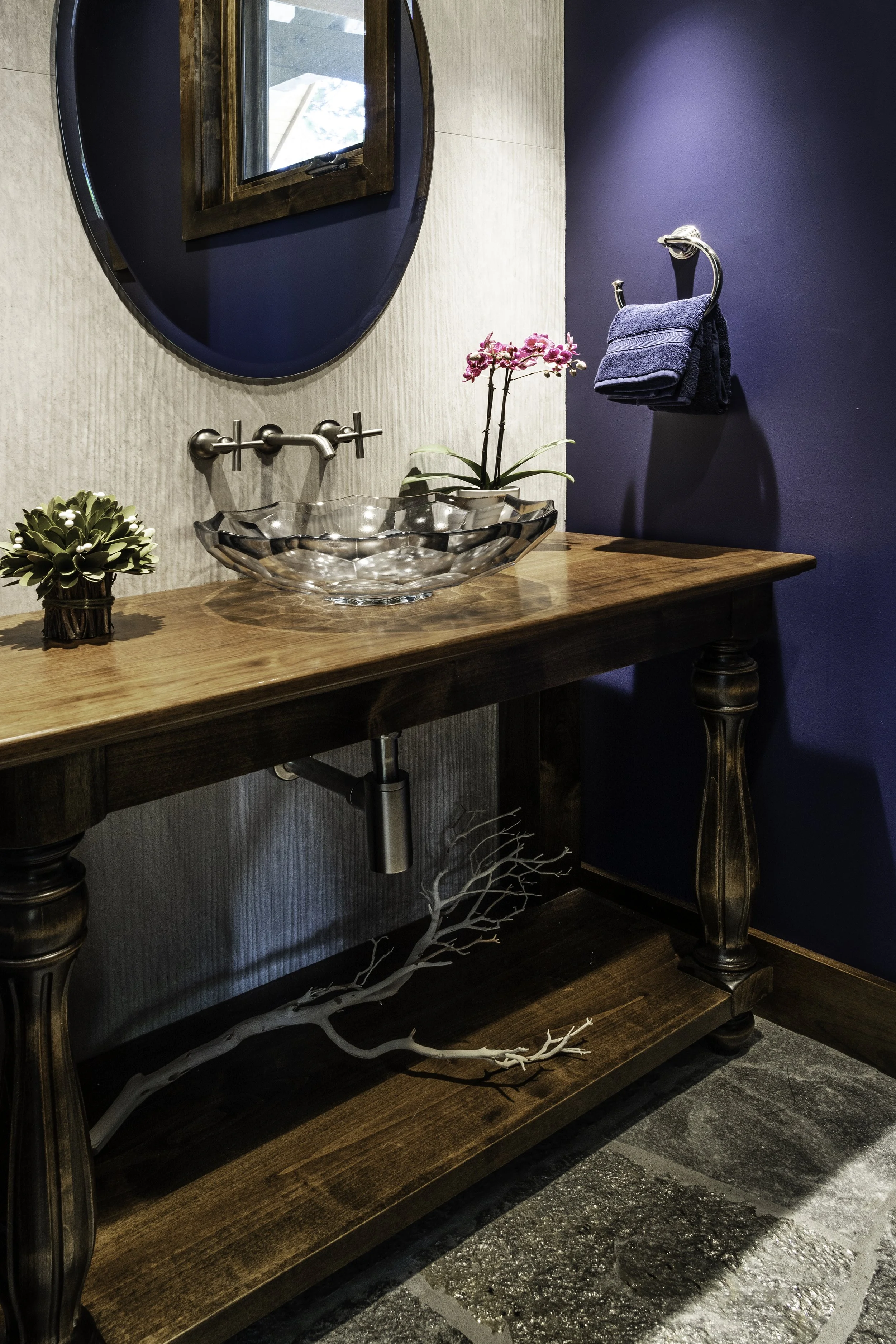 Bathroom vanity with a wooden countertop, glass vessel sink, wall-mounted faucet, pink orchid, small succulent, blue towel, mirror, and decorative branch below the sink.