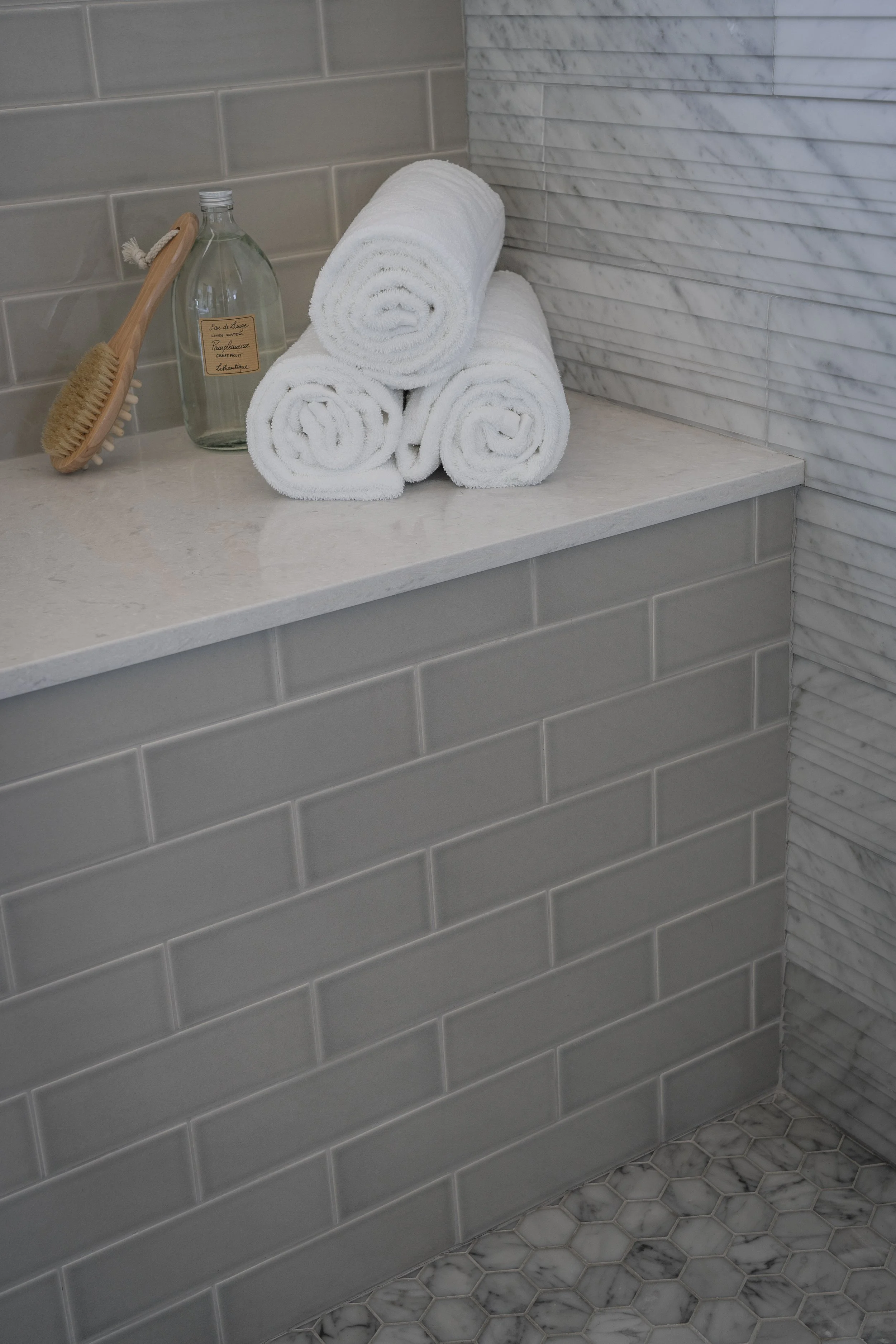 Stacked white towels on a bathroom countertop with a brush, bottle of liquid soap, and marble tile walls.