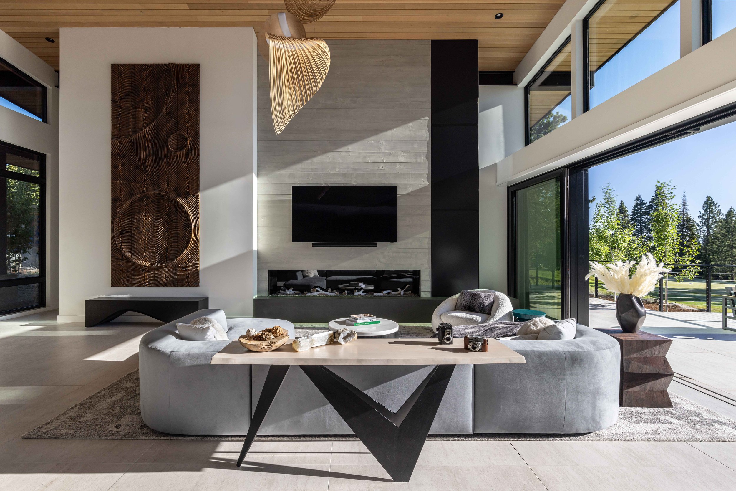 Modern living room with gray sofa, round chairs, a black fireplace, large glass windows, and a view of greenery outside