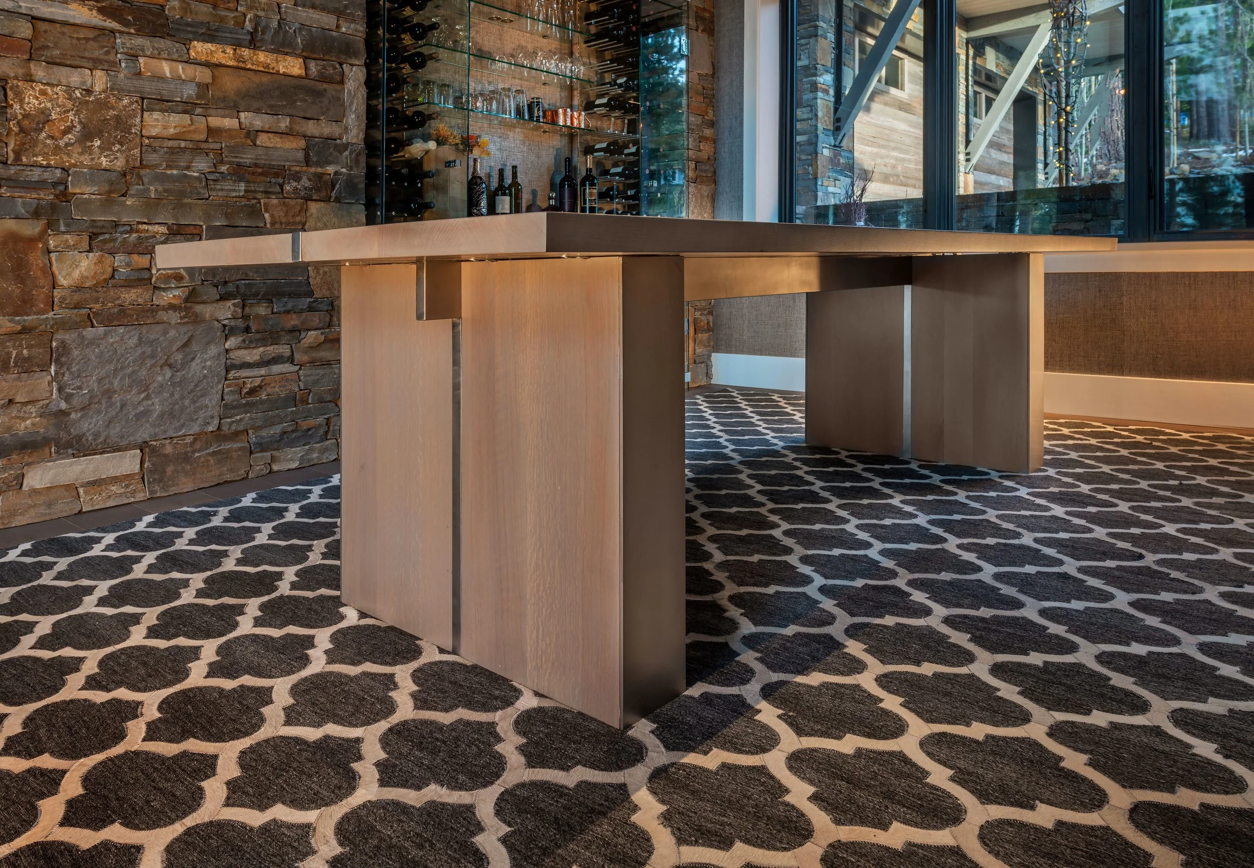 Modern dining table with wooden top and metal legs in a room with a stone accent wall, patterned black and white carpet, and large glass window with a view of trees outside.