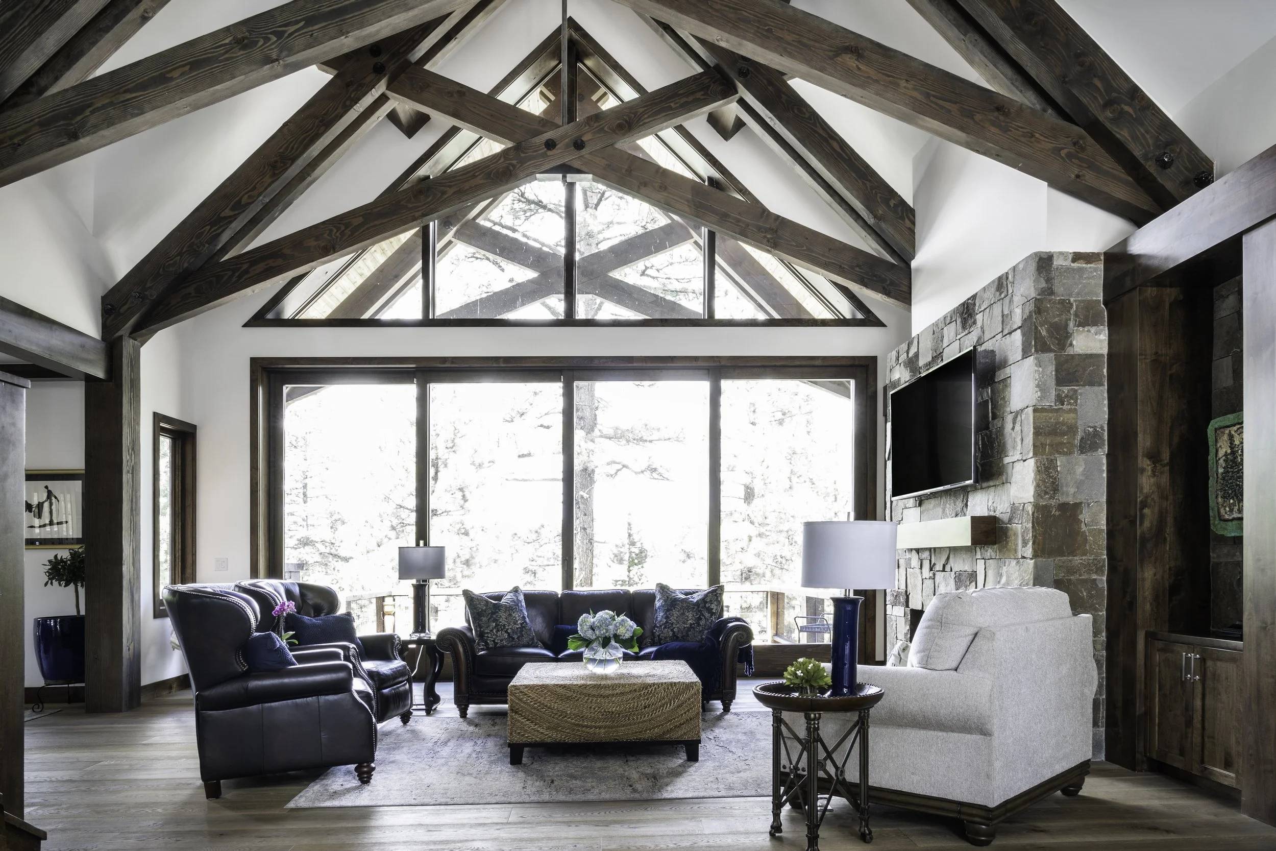 Living room with high vaulted ceiling and wooden beams, large window, stone fireplace with mounted TV, leather and fabric sofas, side tables with lamps, and decorative plants.
